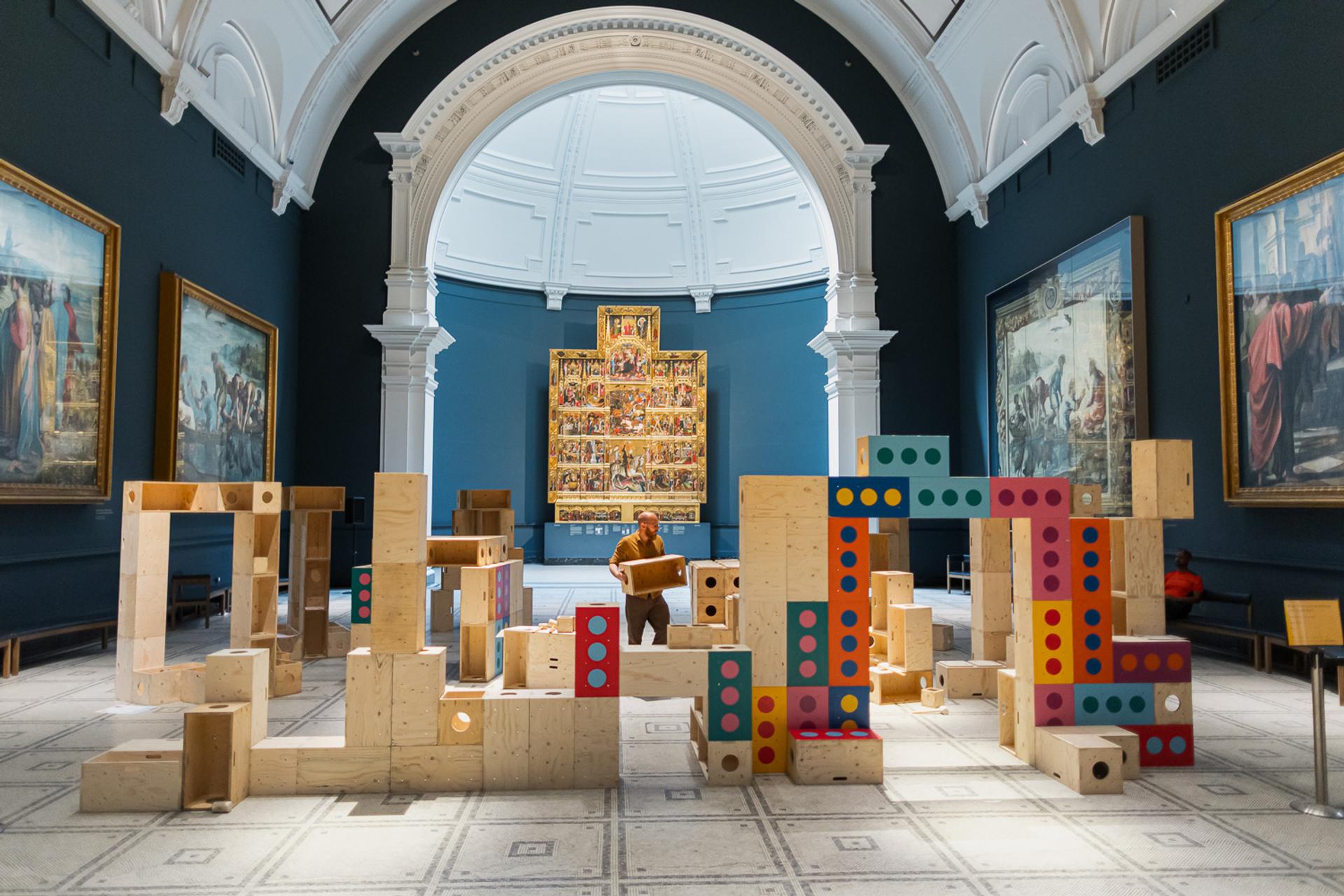 A person stands among large wooden blocks with colourful dots, arranged as a playful installation in the grand V&A museum gallery. Surrounded by classical paintings and an arched ceiling, the scene invites visitors to explore how creativity and upstart ideas can build structures for activism within this iconic space.
