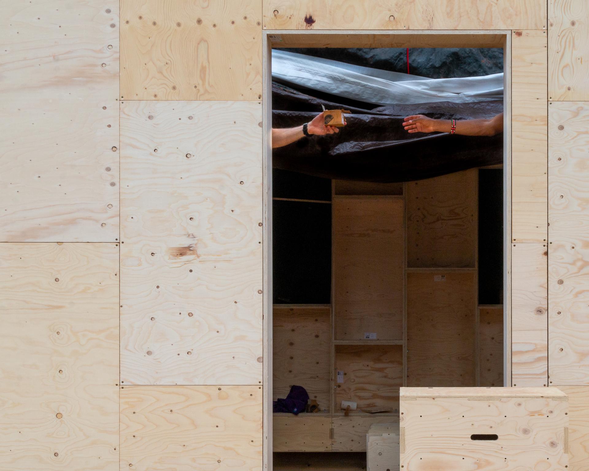 A doorway in a light plywood wall reveals two outstretched arms inside, one passing a small, sustainably sourced wooden block to the other. The room has unfinished wooden shelves and black material above.