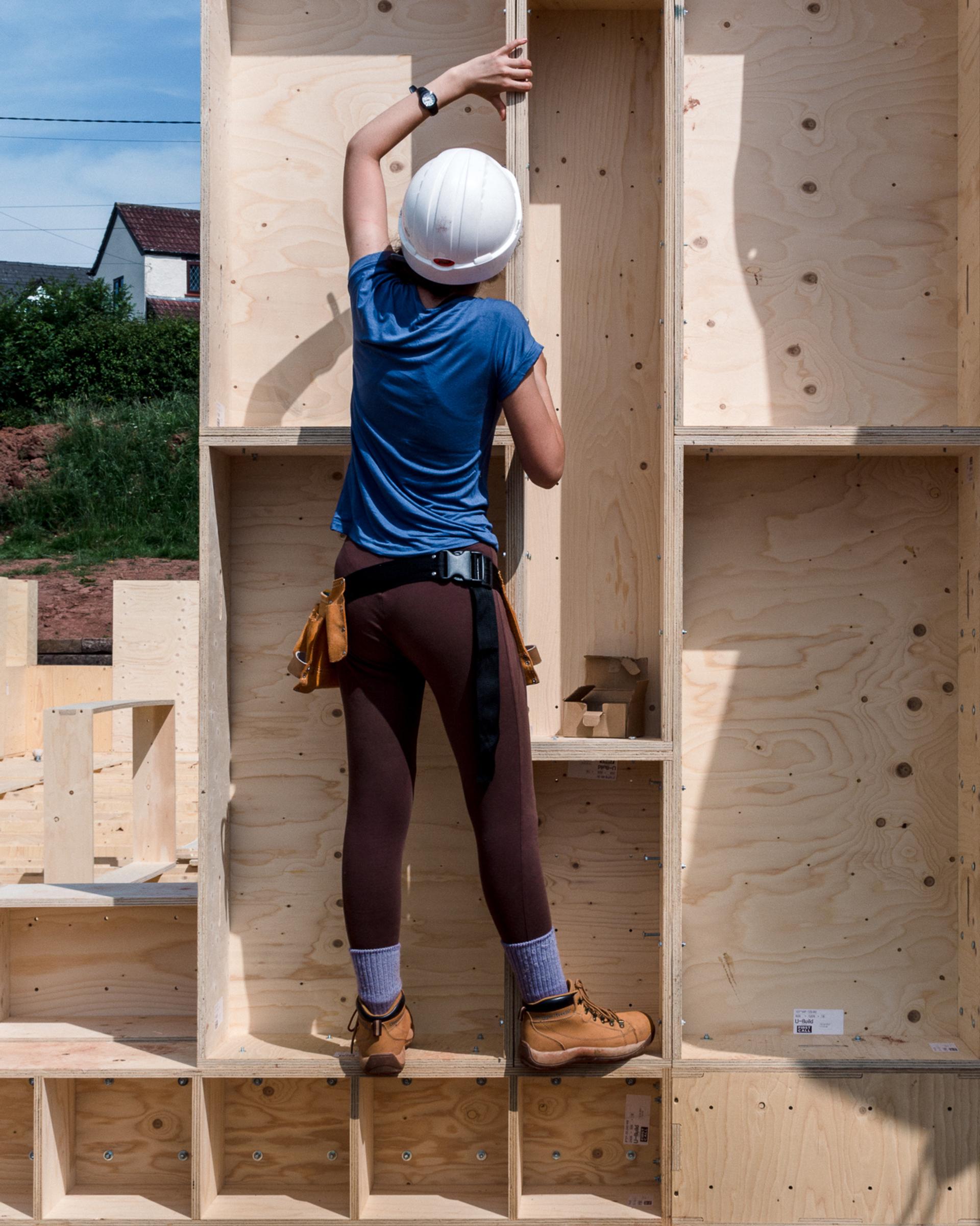 A person wearing a white hard hat, blue shirt, brown trousers, and work boots installs a sustainably sourced wooden panel at a construction site. They are standing on a lower beam and reaching up to secure the tall frame.