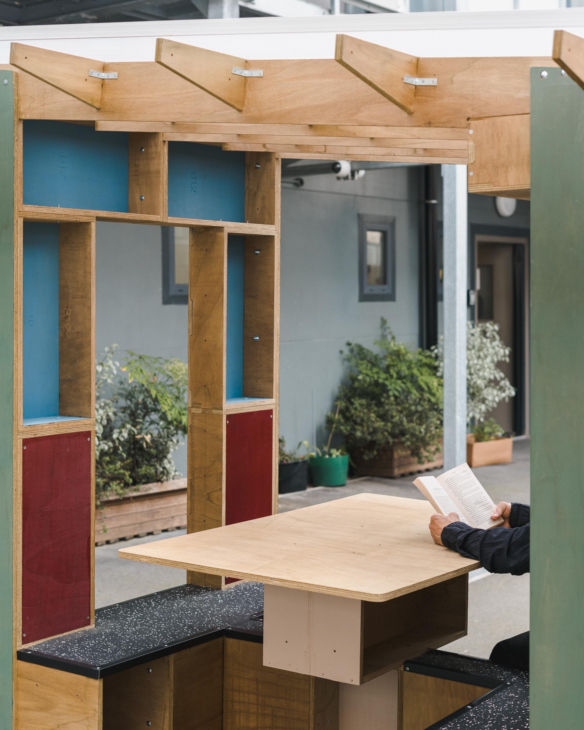 A person sits at a wooden outdoor table with colourful upcycled shelves, reading a book. The scene is set in a courtyard with potted plants and modern building façades in the background.