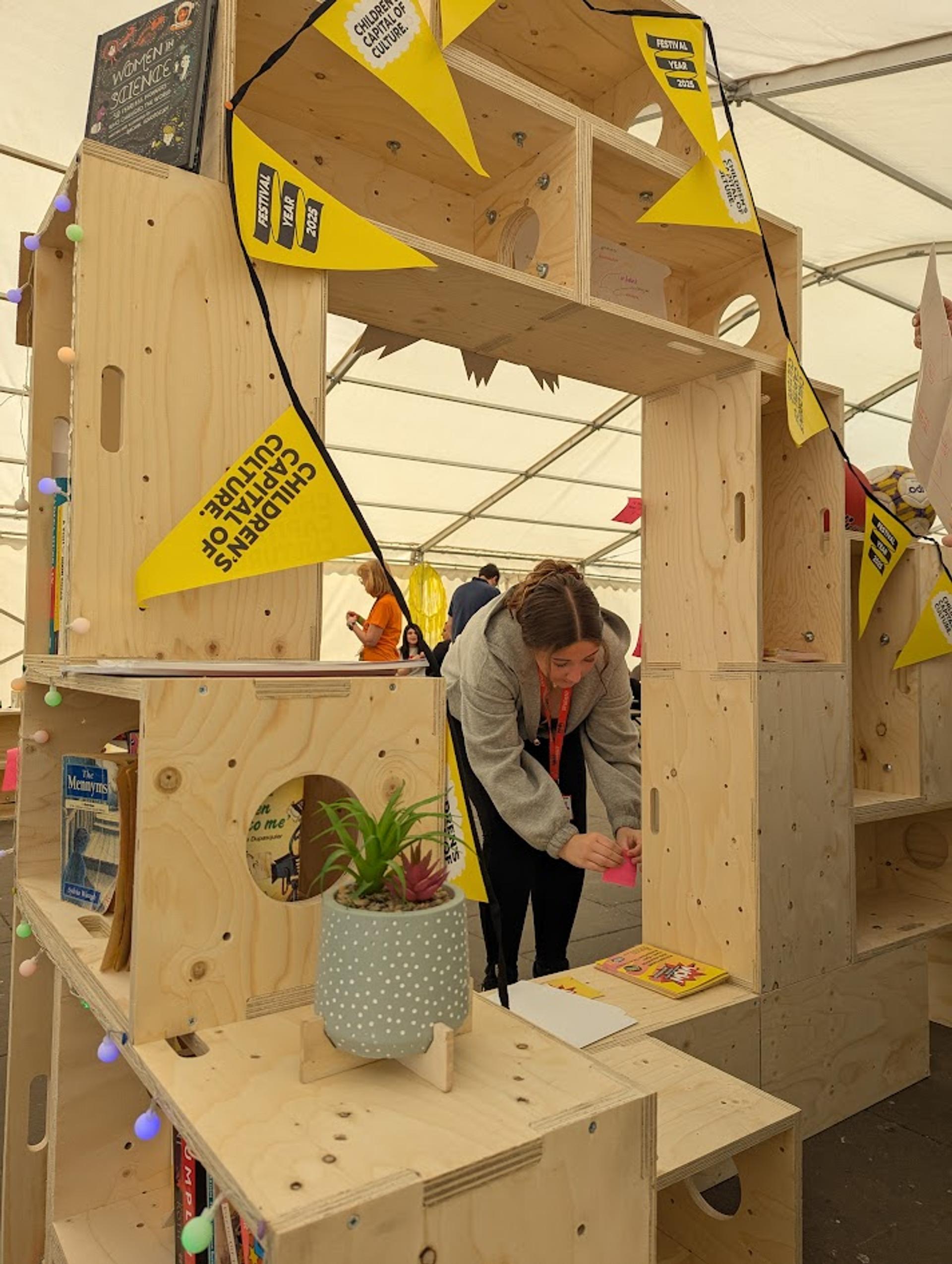 A woman bends over a birch plywood bookcase decorated with yellow bunting that reads Children’s Capital of Culture. Books and a potted plant sit on the sustainable shelves, and she appears to be writing or drawing on a piece of paper.