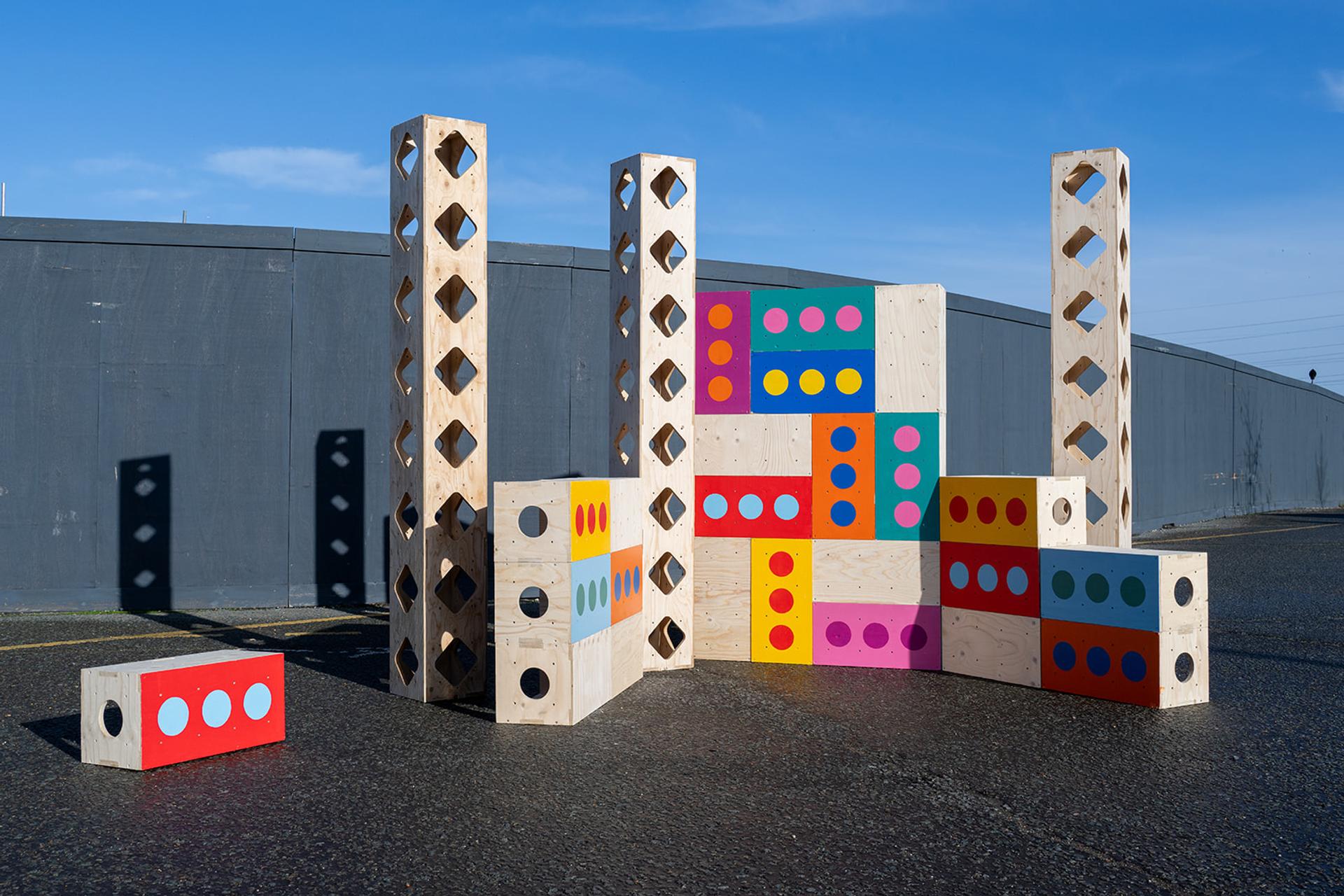 Large wooden blocks, part of the Capturing Box Library, some with coloured circles and others with cut-out holes, are arranged upright and stacked in an outdoor space against a tall grey wall under a clear blue sky.