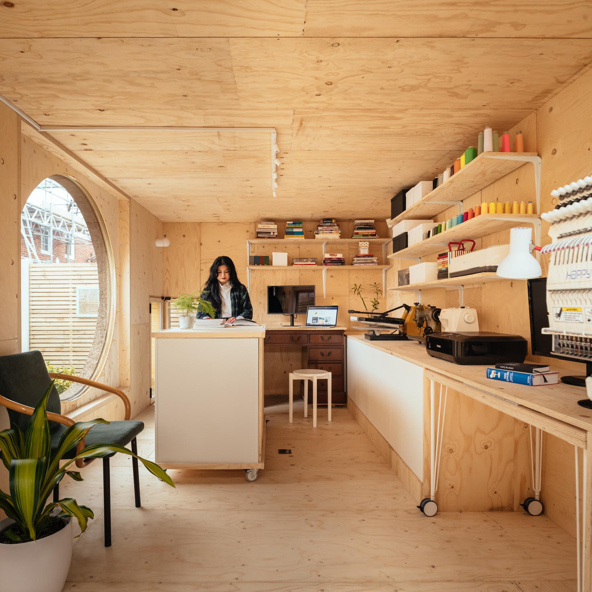 A woman stands at a worktable in a modern, cosy birch studio with shelves of supplies, a laptop, books, and an embroidery machine. Large windows let in natural light whilst a potted plant decorates the modular space.