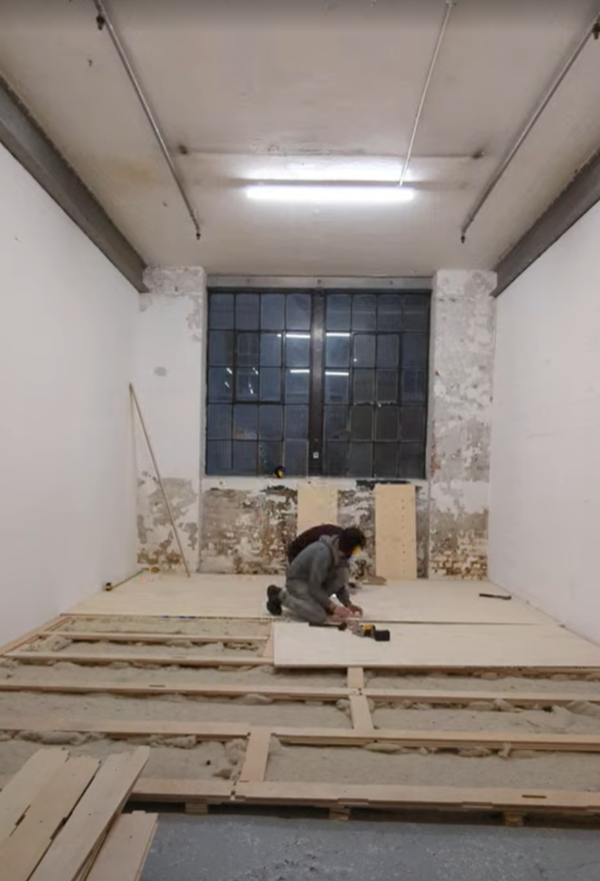 A person kneels on a wooden frame while installing sustainable floorboards in a large, unfinished room with white walls, exposed brick, and a high ceiling with fluorescent lights.