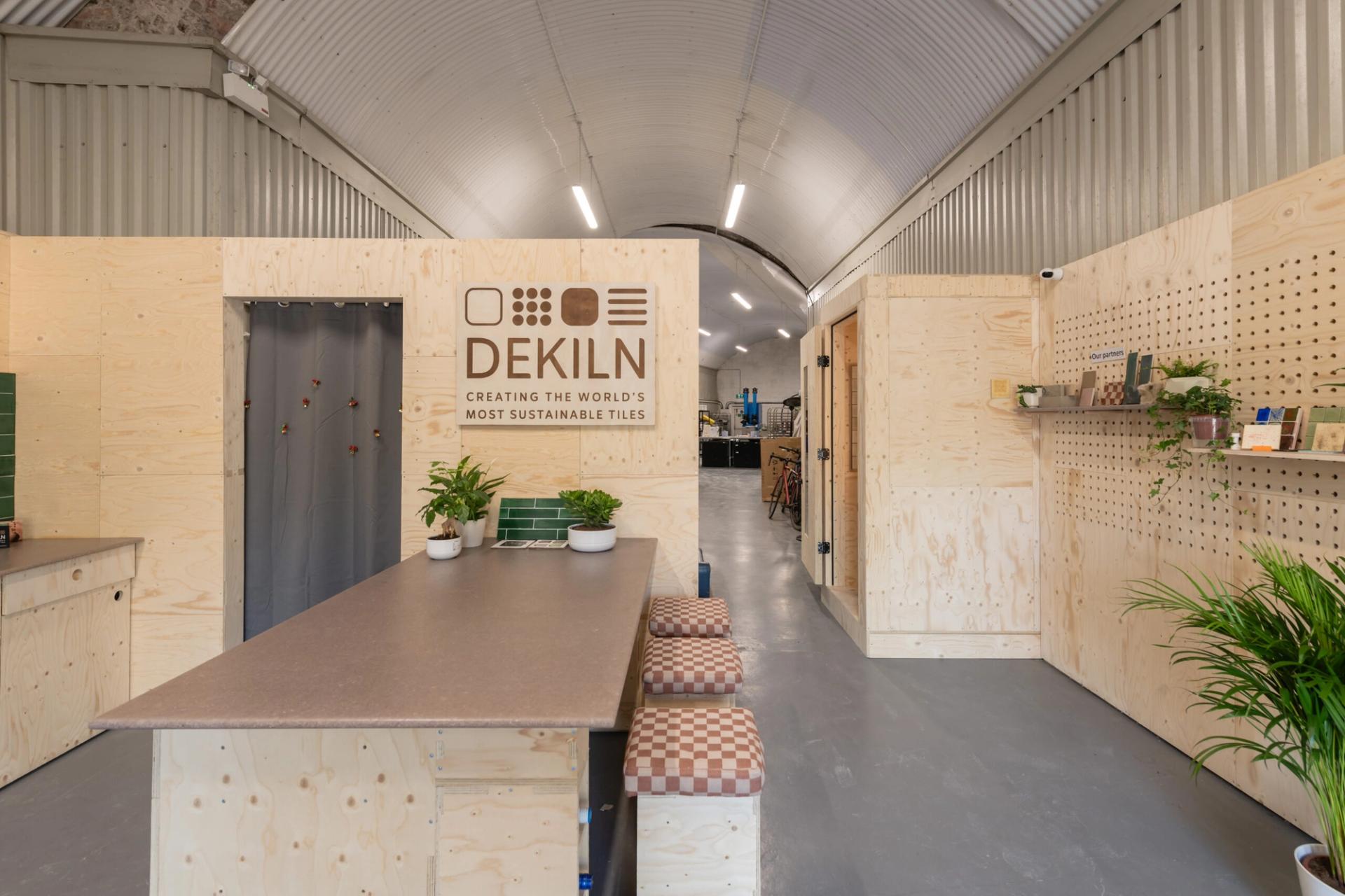 A modern, minimalist Dekiln Showroom with light wood walls, plants, stools around a central table, and a sign reading “DEKILN Creating the world’s most sustainable tiles.” Shelves and a green tile display complete the space.