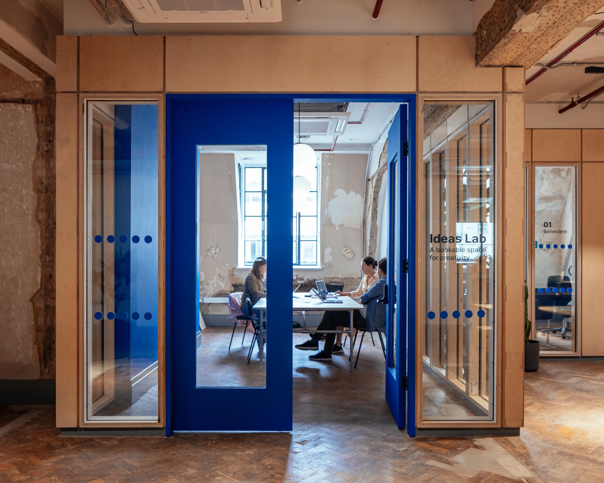 Two people sit at a table working on laptops in the modern Ideas Lab, featuring blue framed glass doors and a large window. The space highlights sustainable timber construction with spruce elements and abundant natural light.
