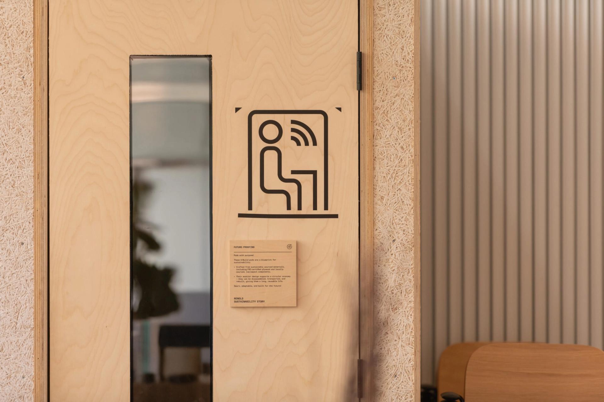 A light wooden door with a narrow vertical window and a black icon showing a person sitting and sound waves, likely at Sustainable Ventures in Manchester. Below the icon is a small plaque. The right side shows part of a corrugated wall and a chair.