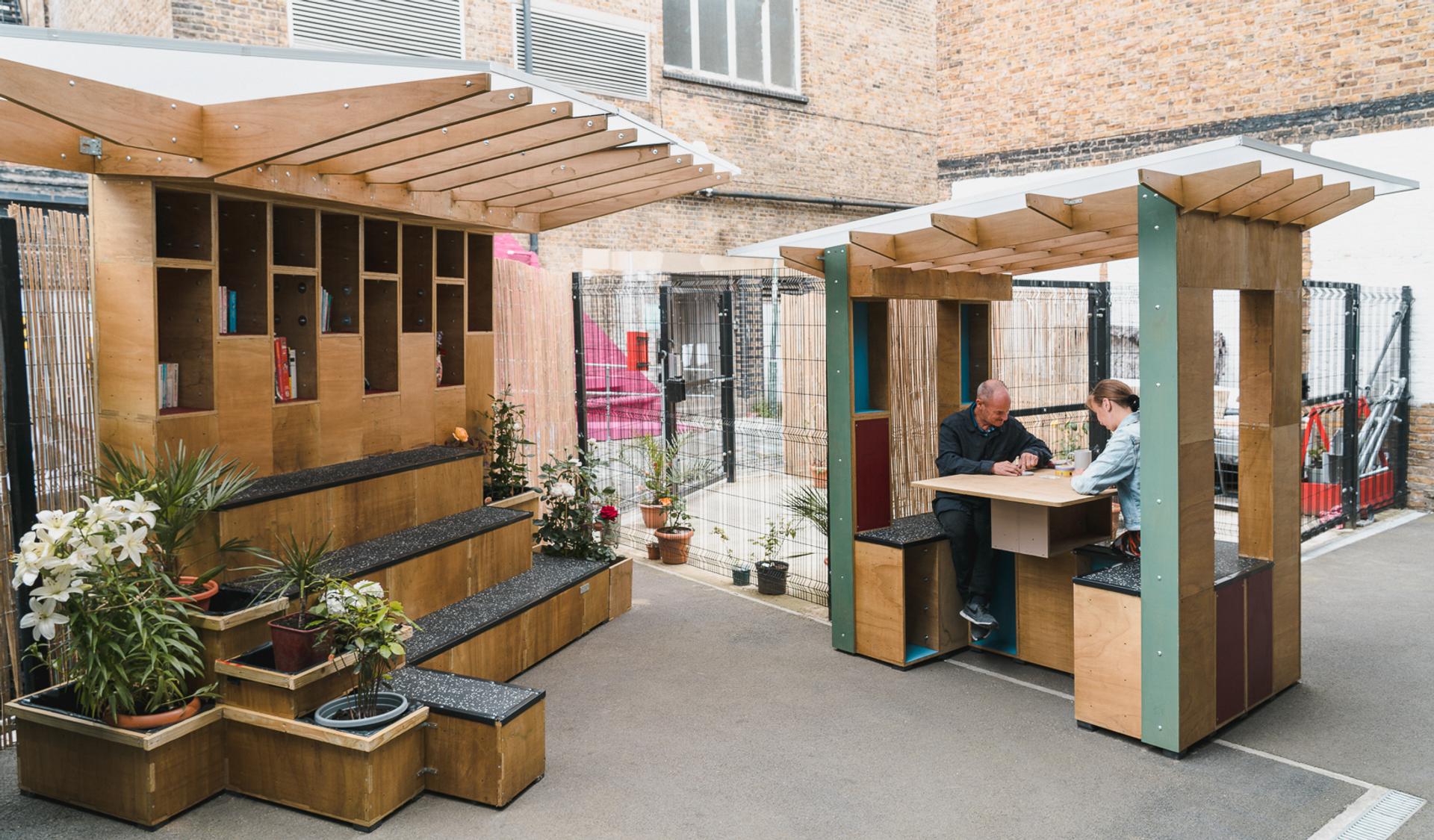 Two people sit at a wooden outdoor table with a pergola roof, surrounded by potted plants and modern wooden structures crafted from sustainably sourced materials, in a fenced urban courtyard with brick and white walls.