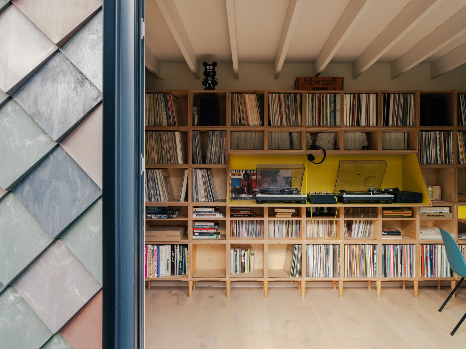 A cosy Record Room in East London with wooden shelves filled with vinyl records, a yellow record player, speakers, and stylish decor. The neatly organised shelves sit beside an exterior wall with grey-green tiles on the left.