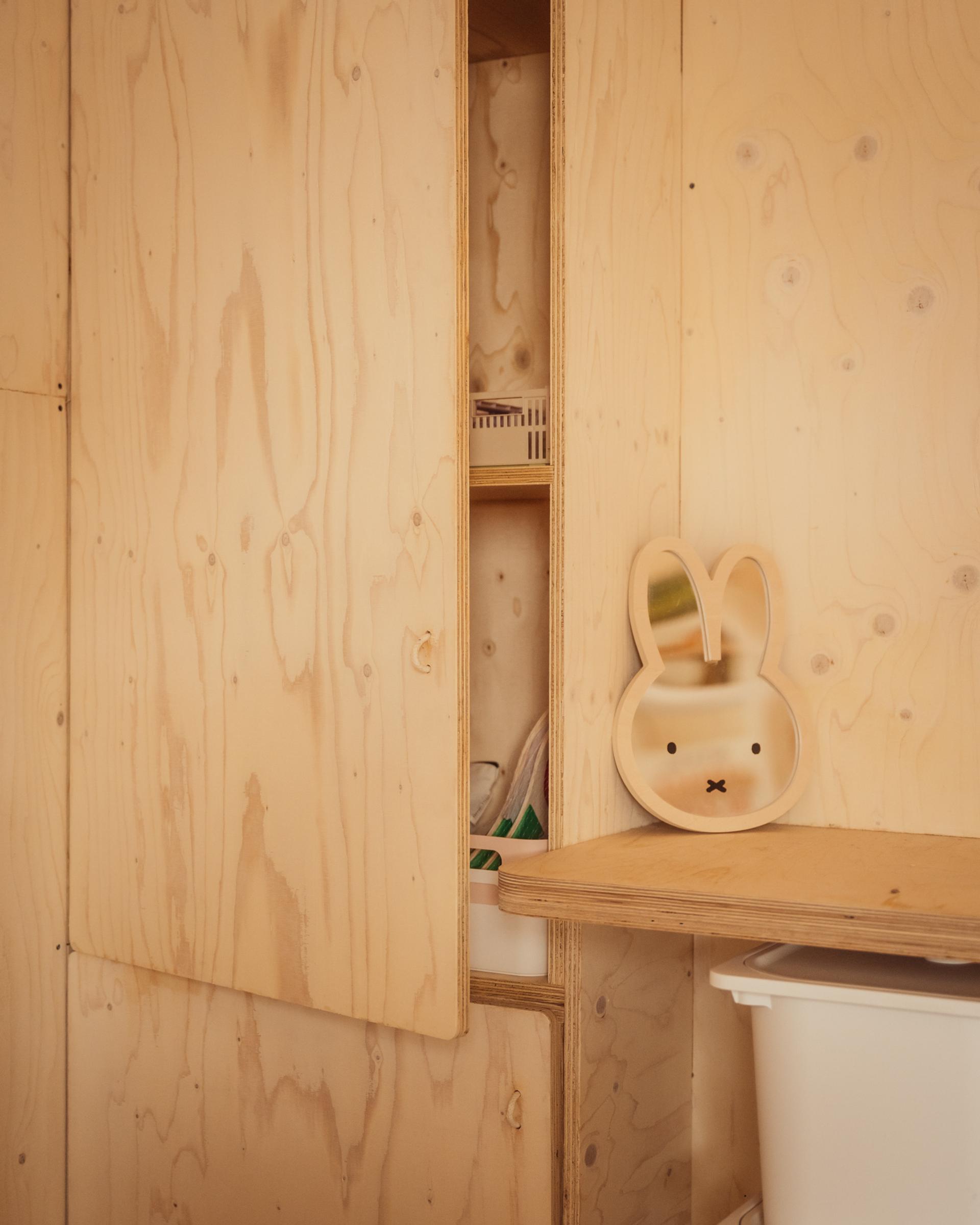 A wooden cupboard with an open door revealing shelves sits next to a rabbit-shaped mirror on a desk, capturing the thoughtful architectural design of this home extension.