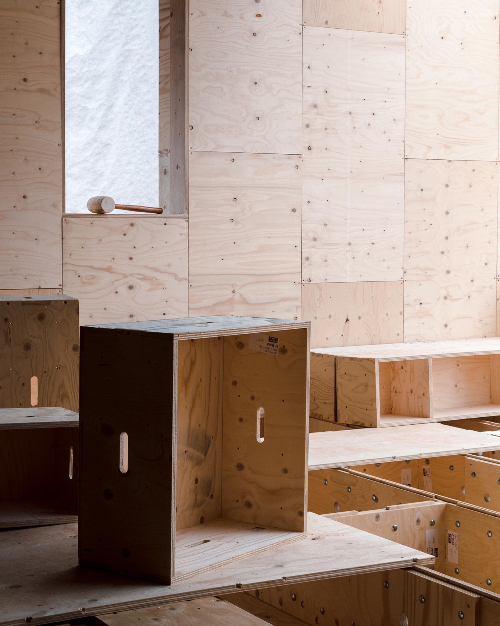 A room with walls, floors, and steps made of sustainably sourced, light-coloured plywood. A wooden box with handle cut-outs stands upright in the centre. Sunlight enters through a tall narrow window covered by a sheer white curtain.