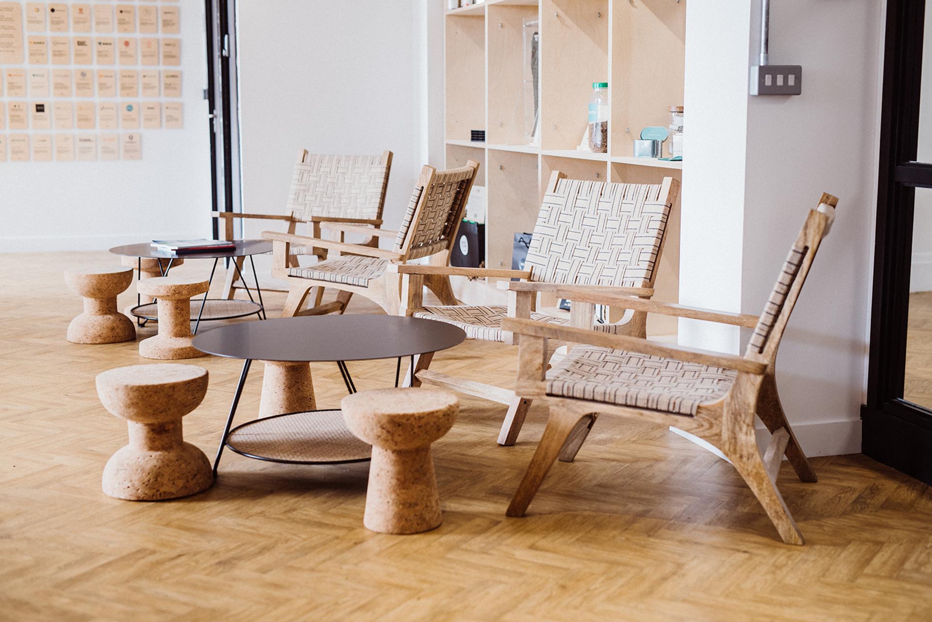 A modern lounge area with spruce wooden armchairs, modular round cork stools, and a circular coffee table on a patterned wood floor. Shelves and a wall with notes are visible in the background.