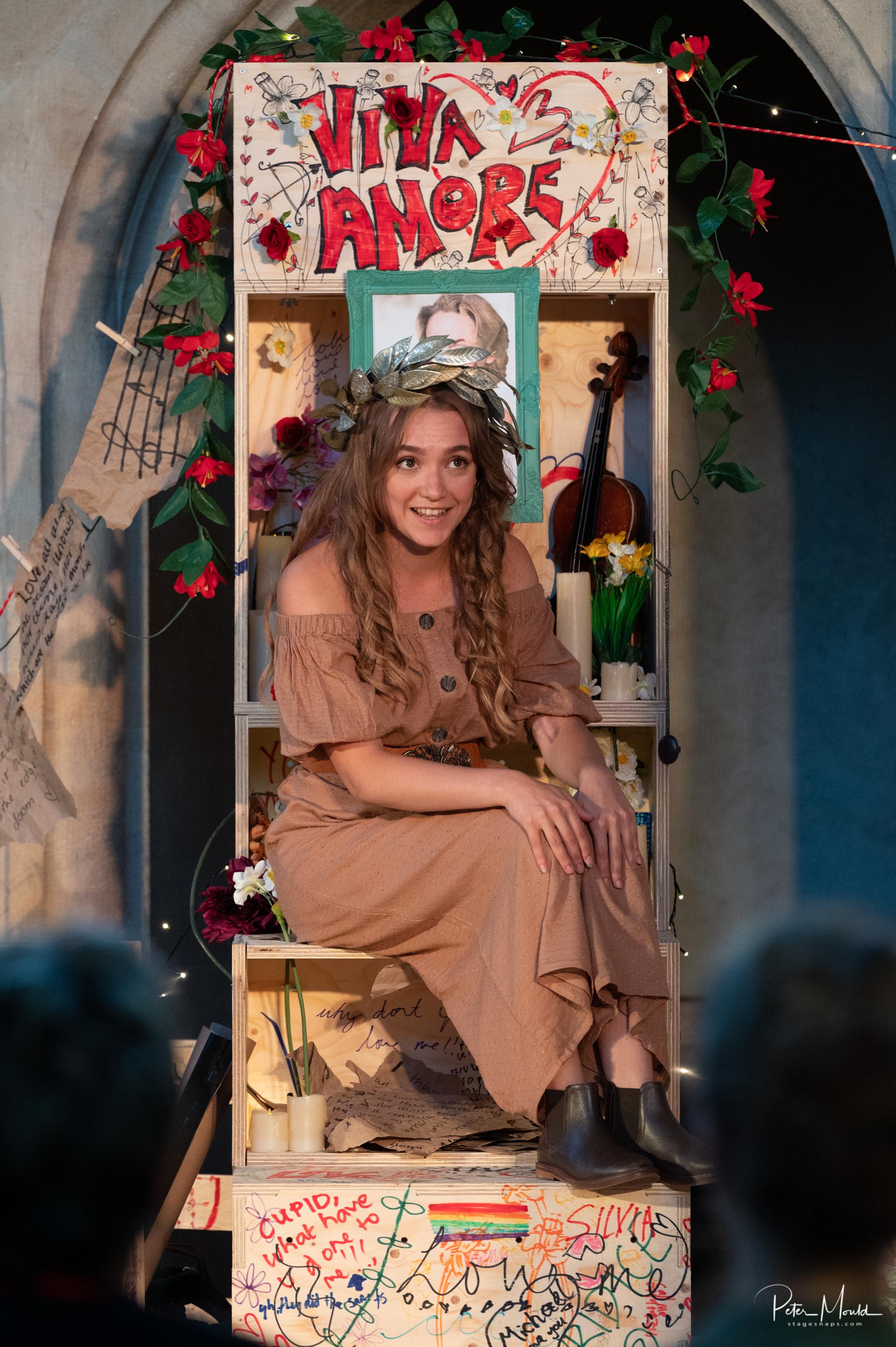 A woman in a brown dress and floral crown sits on a modular wooden chair crafted from plywood, with “Viva Amore” above her. Surrounded by flowers, notes, and fairy lights, she smiles and interacts warmly with the audience.