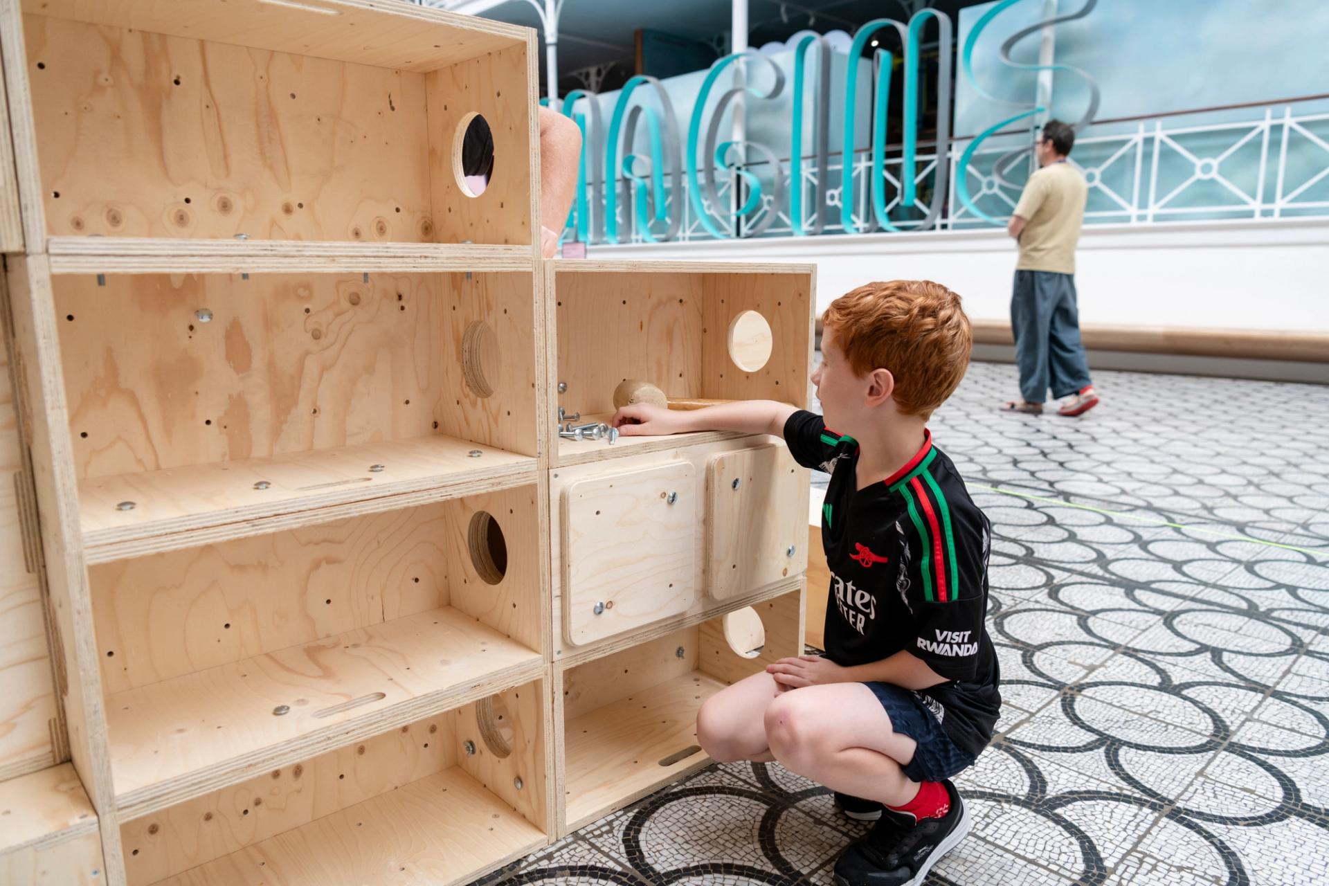 A young boy with red hair kneels on the ground, interacting with a large U-Build wooden structure made of open cubes. He is placing an object inside one of the boxes whilst another person stands in the background.