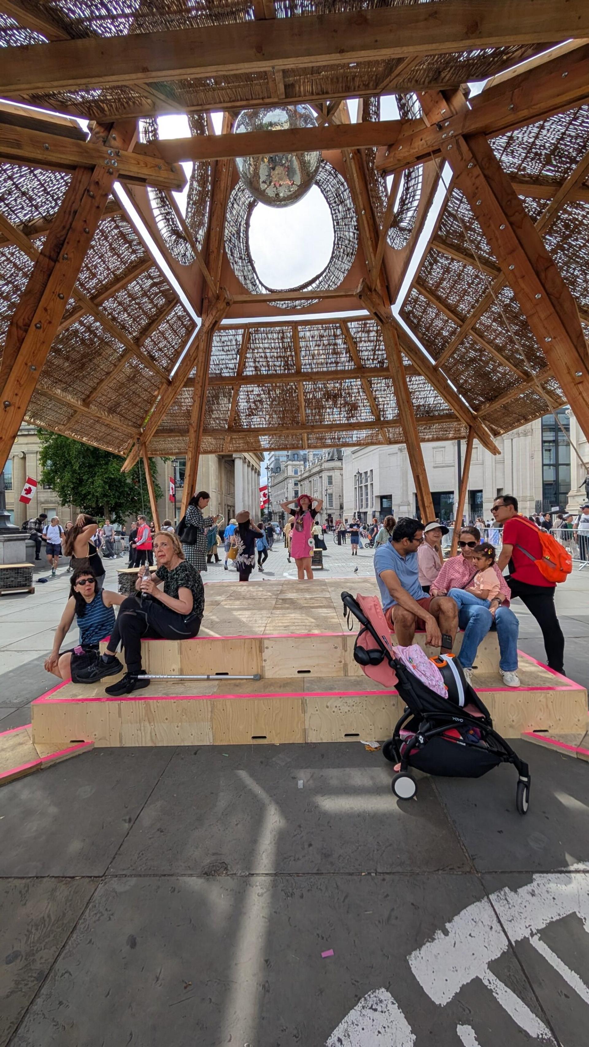 People sit and relax under a large wooden pavilion with a woven roof in a busy urban square near the National Gallery. A pram and coffee cup are in the foreground, whilst others walk and socialise in the background, celebrating the Triumph of Art.