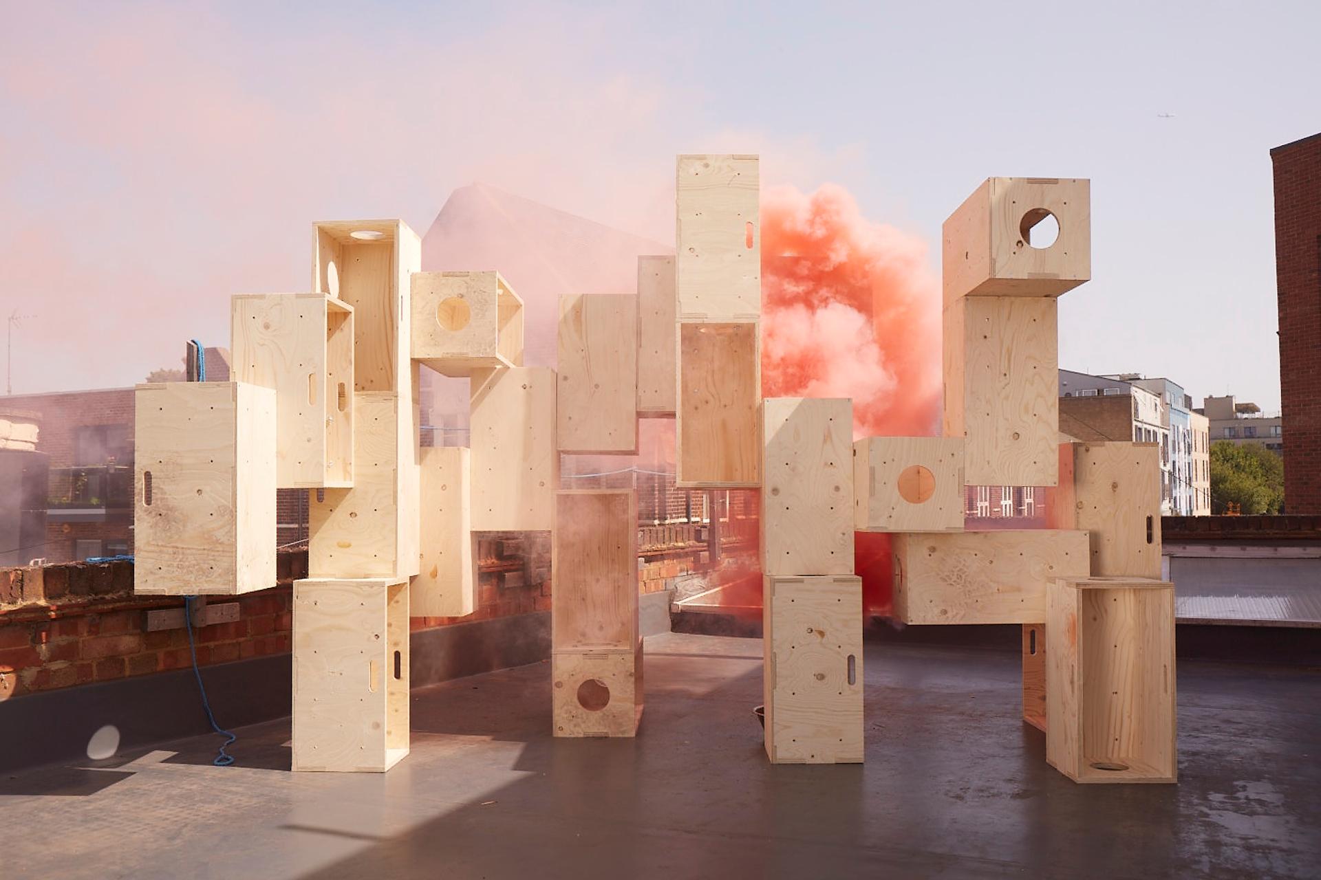 A group of large, light plywood boxes is stacked in an abstract arrangement on a rooftop, with orange smoke billowing behind them and buildings visible in the background under a clear sky.
