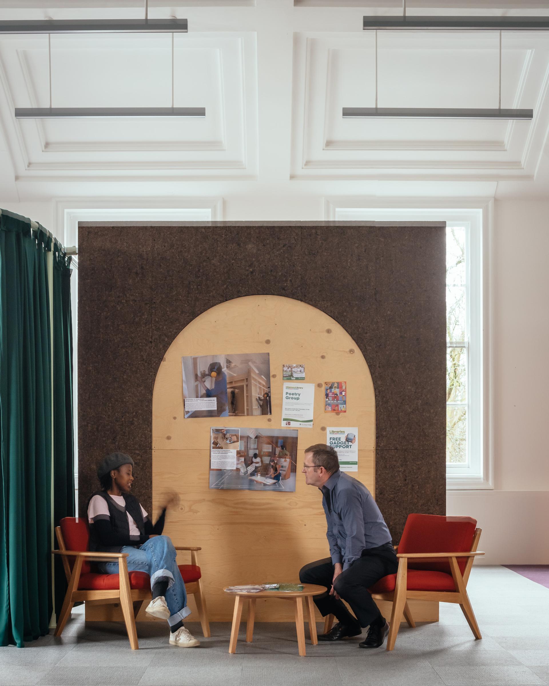 Two people sit on red armchairs facing each other in a modern room, talking. Behind them is a timber construction panel with an arched cut-out, decorated with photos and flyers. A small round table is between them.