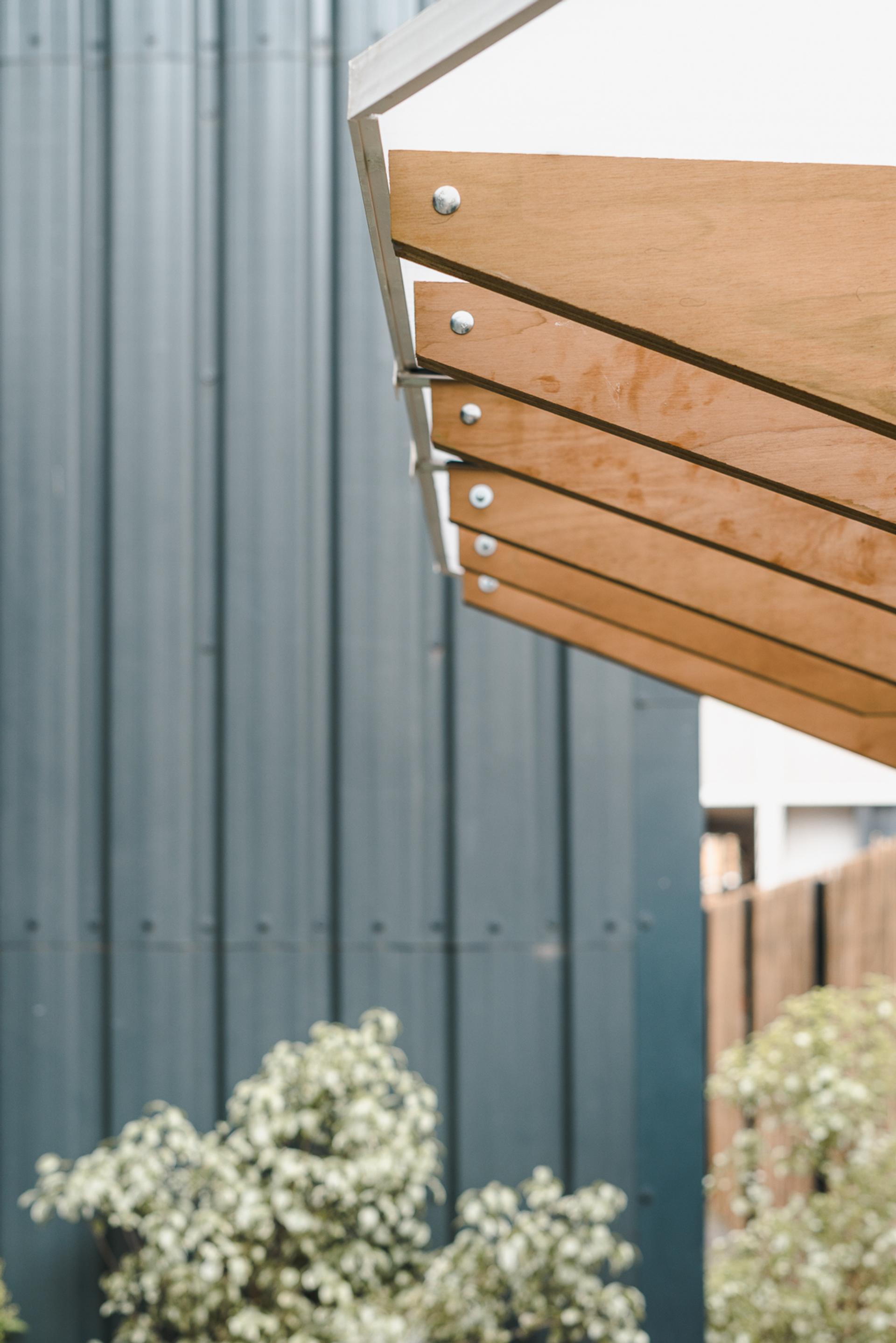 A close-up of a wooden pergola roof with metal fastenings, photographed against a background of a corrugated metal wall and lush green plants below—showcasing smart reuse in line with circular economy principles.