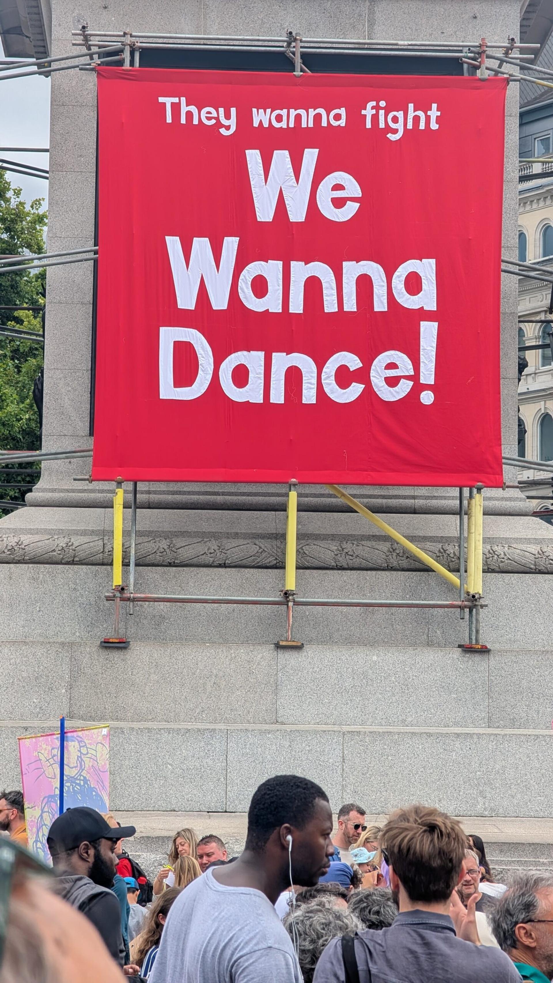A large red banner reads “They wanna fight. We wanna dance!” above a crowd gathered outdoors in front of a stone monument. The lively scene reflects a spirit of unity, echoing the values of recycle and upcycle within a circular economy.
