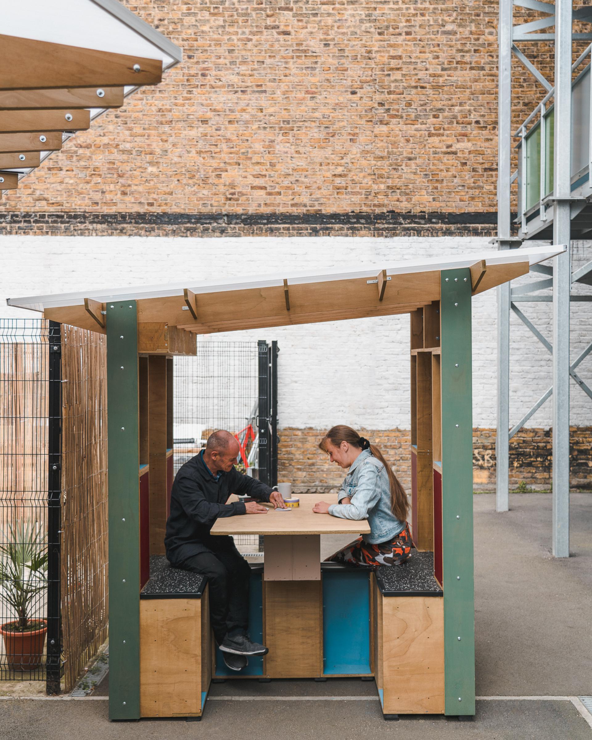 Two people sit opposite each other at a small outdoor wooden table with benches, upcycled from old materials, under a simple shelter near a brick wall and fencing. They appear to be writing or drawing on paper.