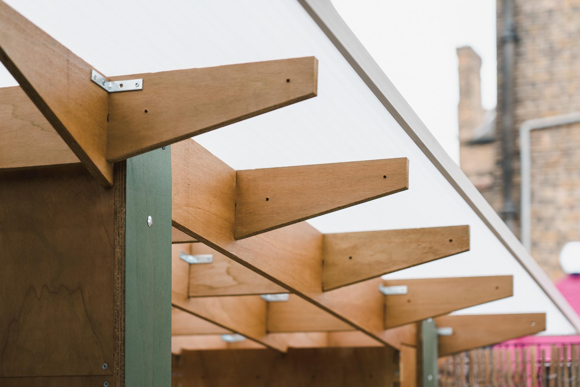 Close-up of a wooden structure with triangular beams supporting a translucent roof, showcasing sustainability against a blurred background with stone buildings.