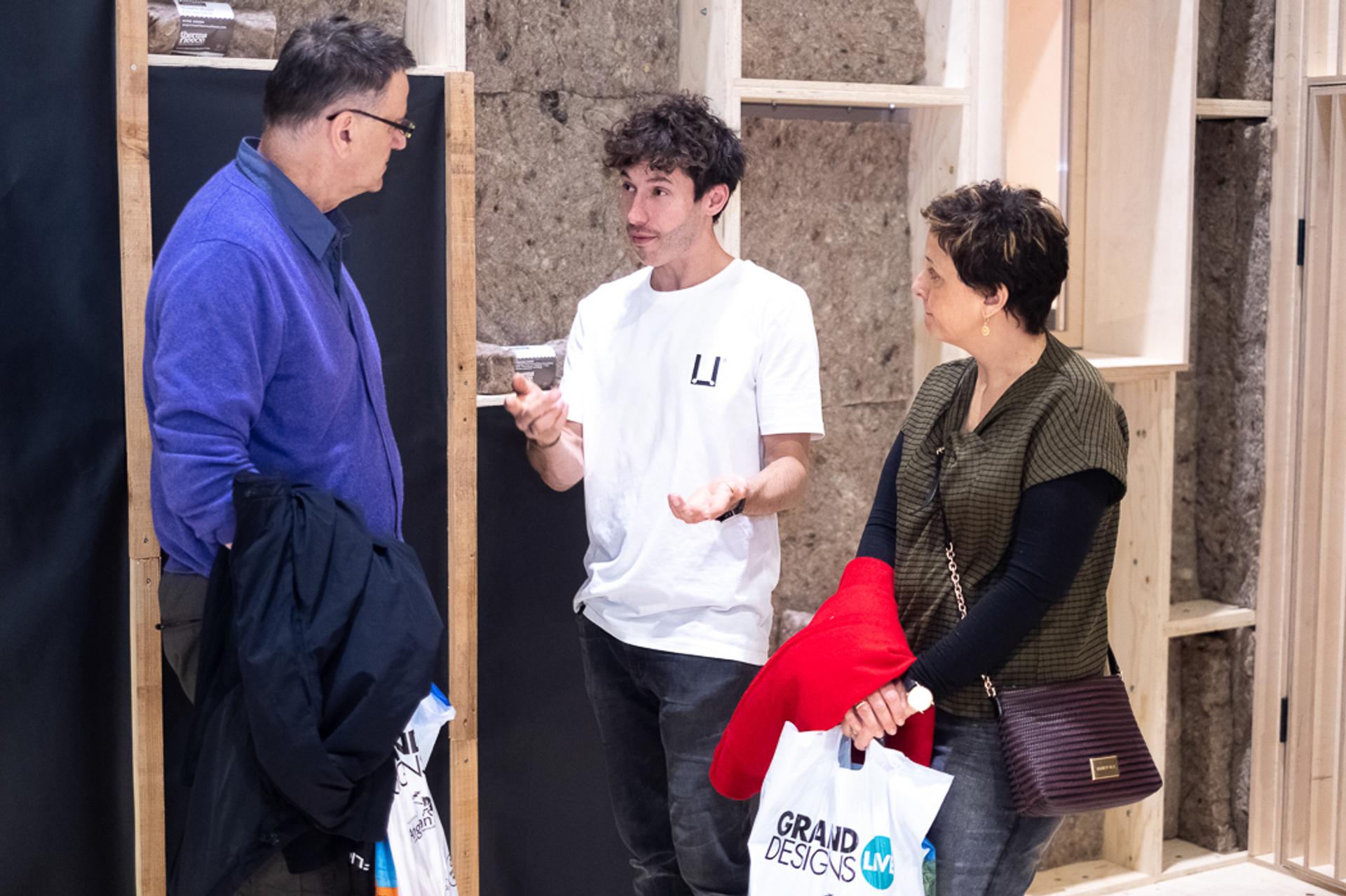 A young man in a white T-shirt talks to a man in a blue jacket and a woman with a red jumper and Grand Designs Live tote bag, standing in front of a partially built Timber Construction wall.