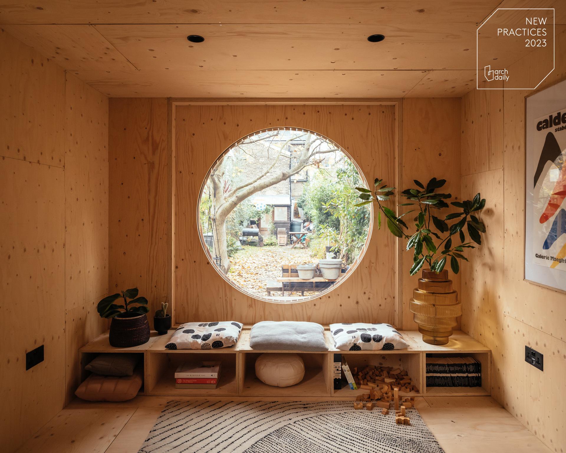 Cosy wooden room with modular built-in bench and cushions beneath a large round window, looking out to a garden with trees and outdoor seating. Spruce shelves hold plants and books, creating a warm, inviting atmosphere.