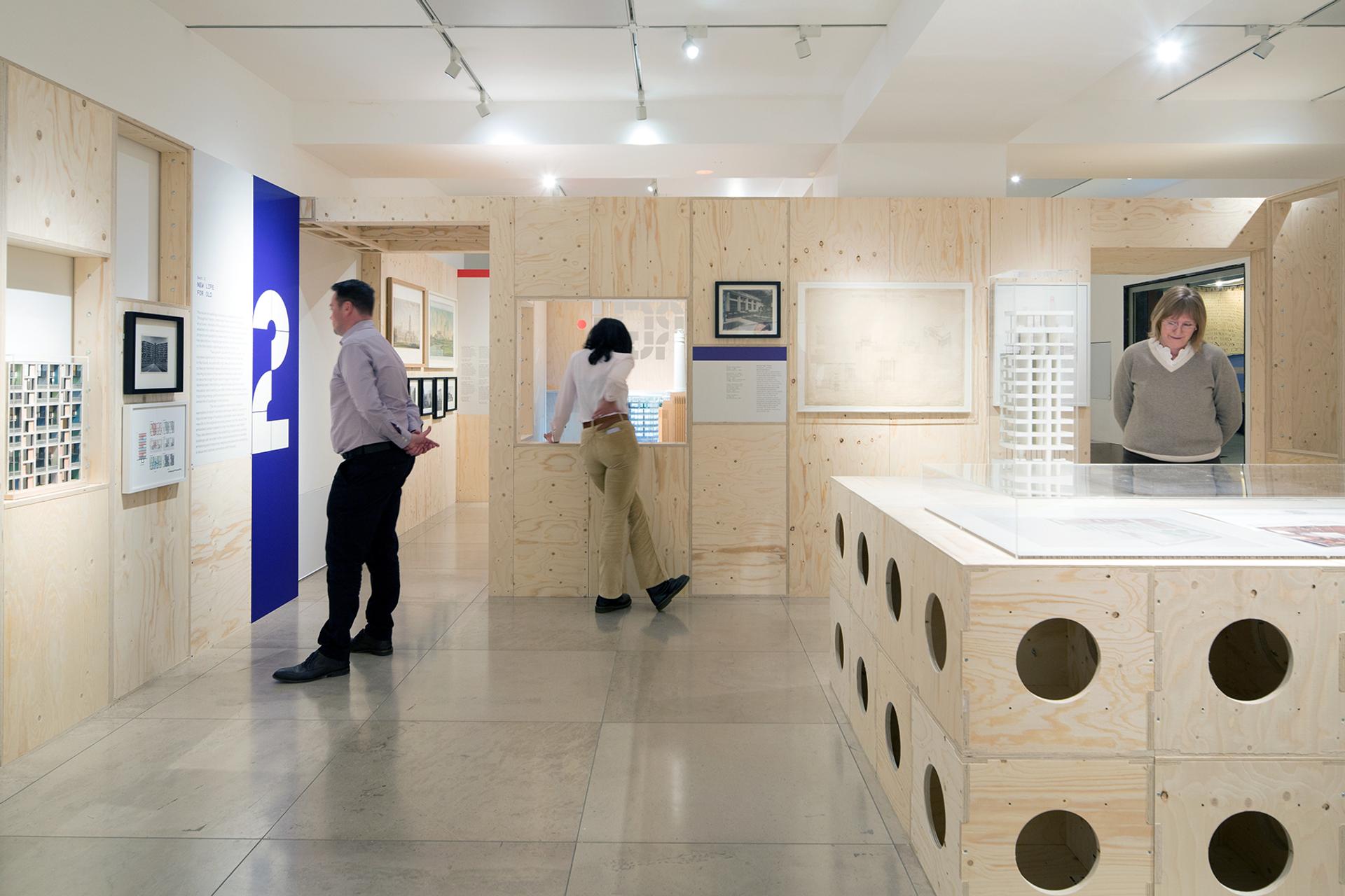 Three people view exhibits in a modern, birch-panelled gallery with framed photos and architectural displays. The polished concrete floor and spotlit ceiling highlight the modular design and showcase innovative timber construction techniques.