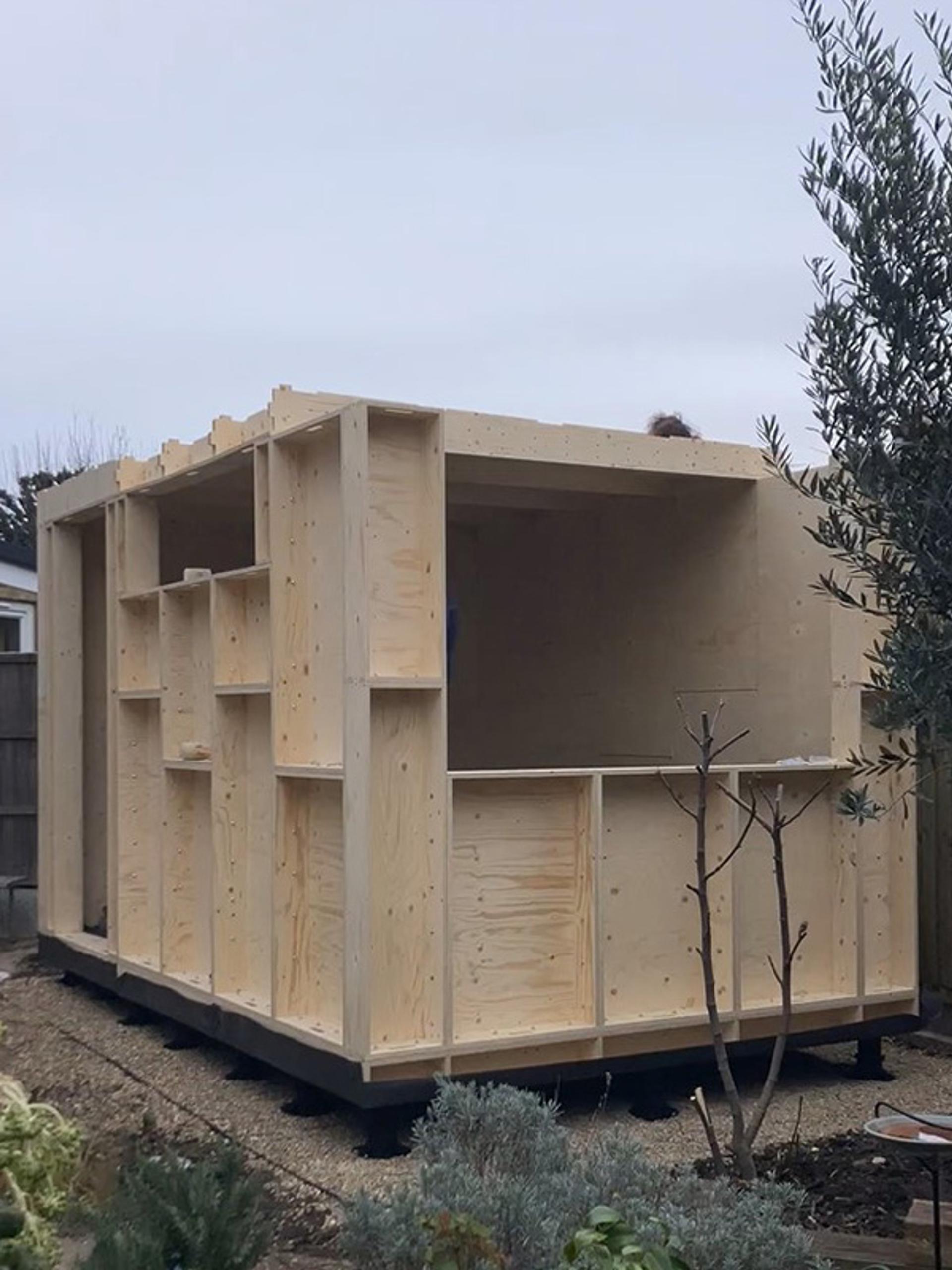 A small timber construction shed or cabin stands unfinished in a garden, with exposed plywood walls and open window frames. Surrounded by plants and a cloudy sky, this modular structure blends naturally with its leafy surroundings.