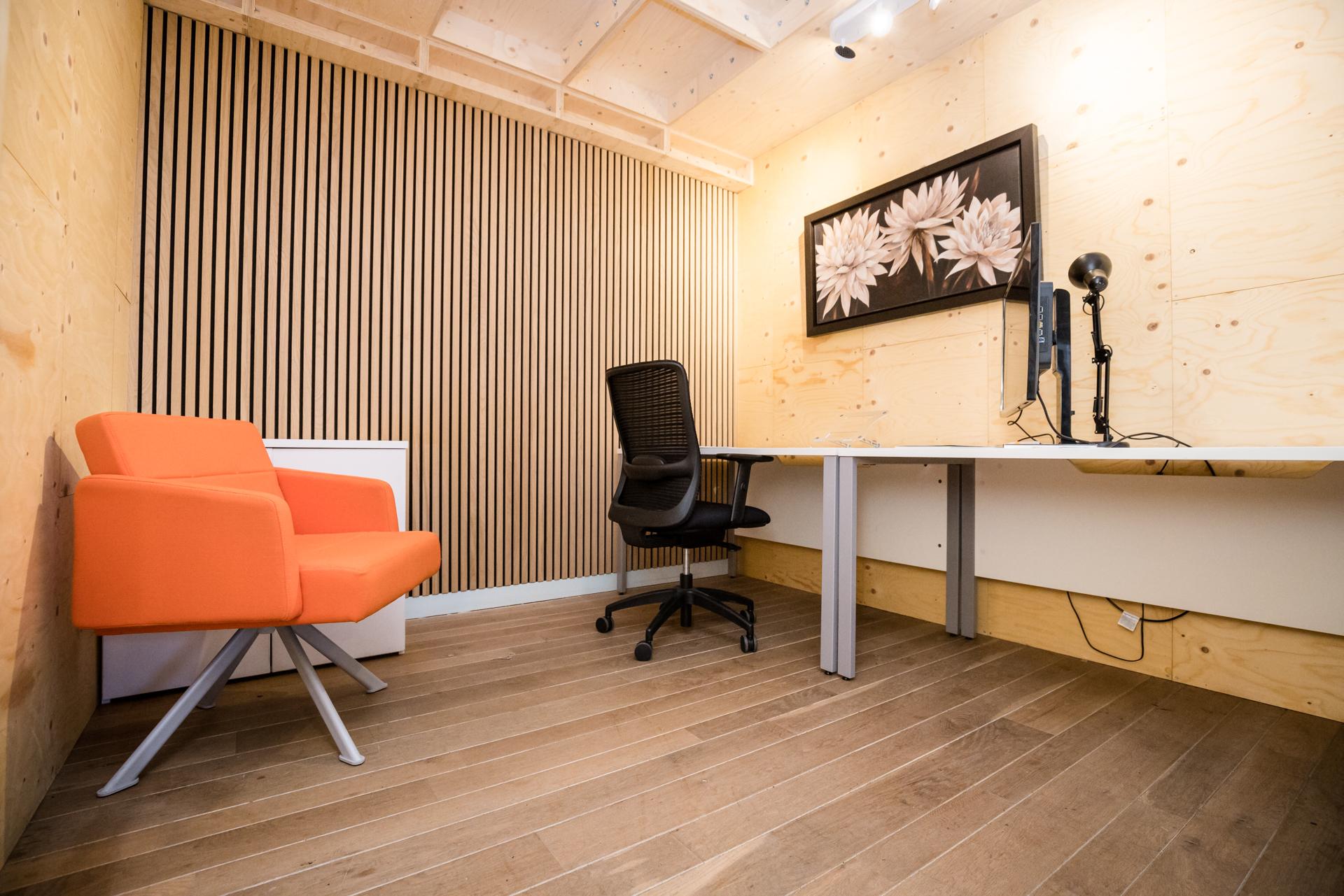 A modern office with birch wooden walls and floor, featuring an orange armchair, a black office chair at a desk with a computer, microphone, and a framed flower artwork on the wall.