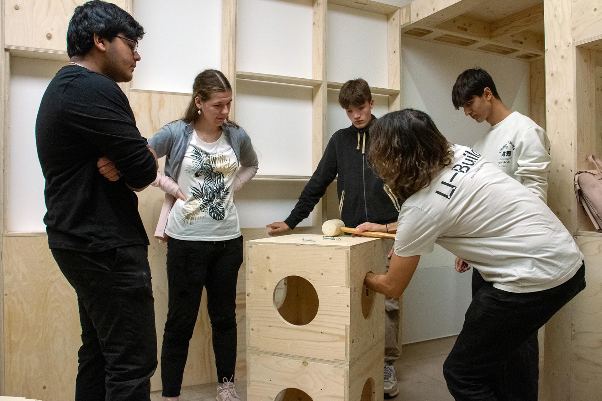 Five young people are gathered in a wooden room, focusing on a sustainable, cube-shaped timber construction with circular openings as one person writes or draws on it while others observe attentively.