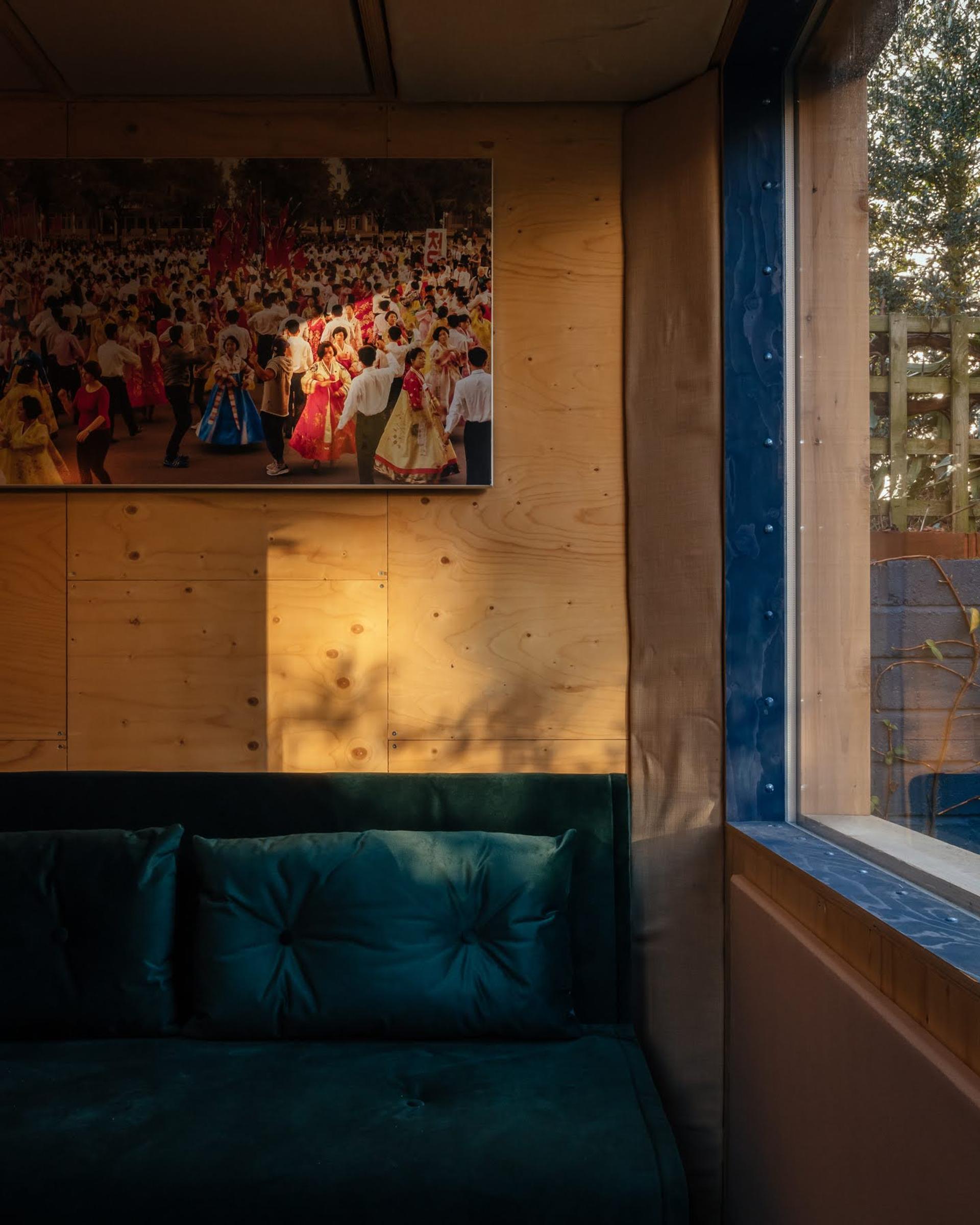 A sunlit room with a green velvet sofa, birch timber construction walls, and a large window. A colourful photograph of people in traditional clothing at a festival hangs above the sofa.