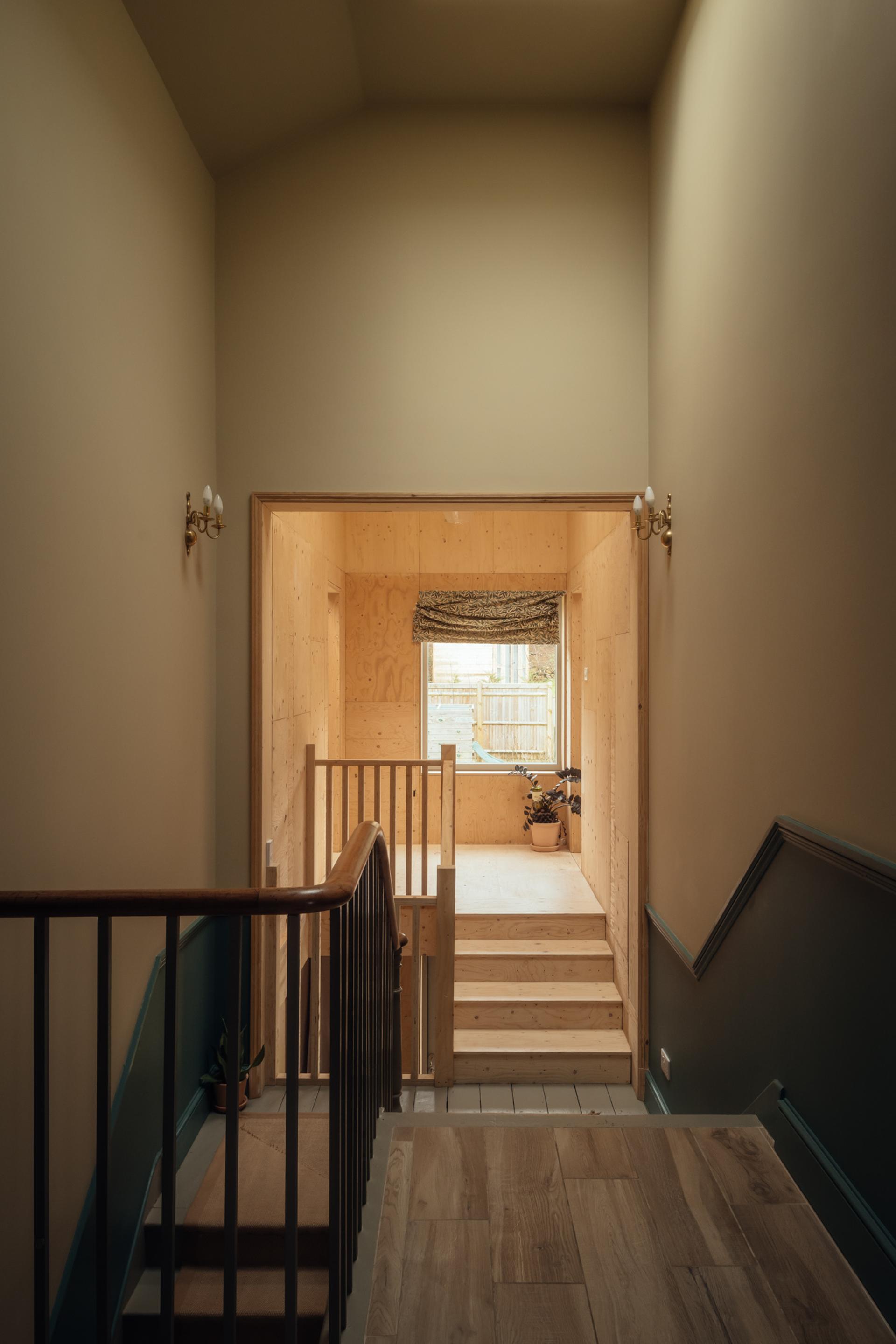 A view from a staircase landing looking into a room with timber cladding and a small set of wooden stairs leading to a window.