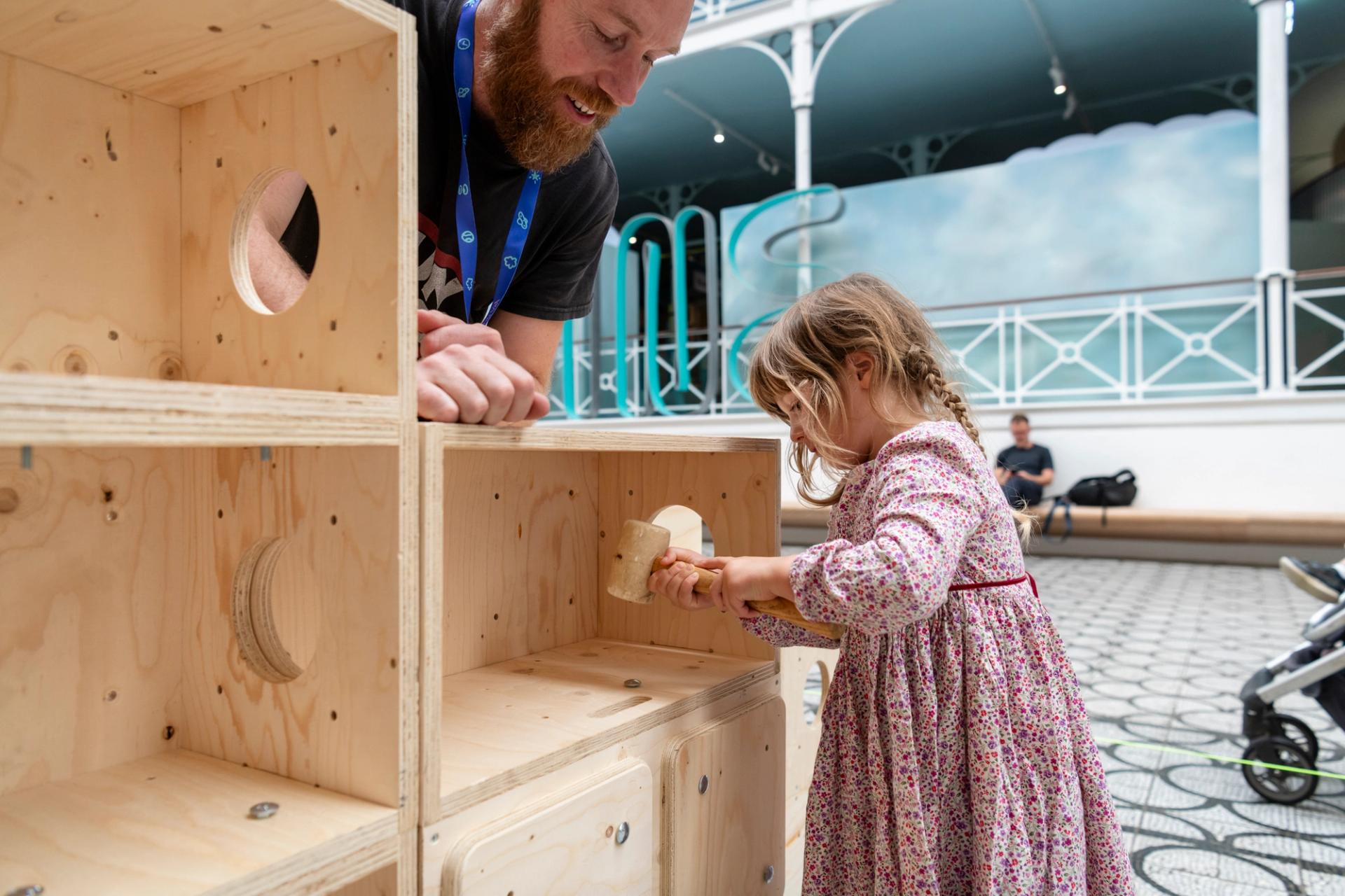 A young girl in a floral dress uses a wooden U-Build tool on a wooden play structure, while an adult smiles and watches her. The scene is set indoors with a patterned floor and people in the background.