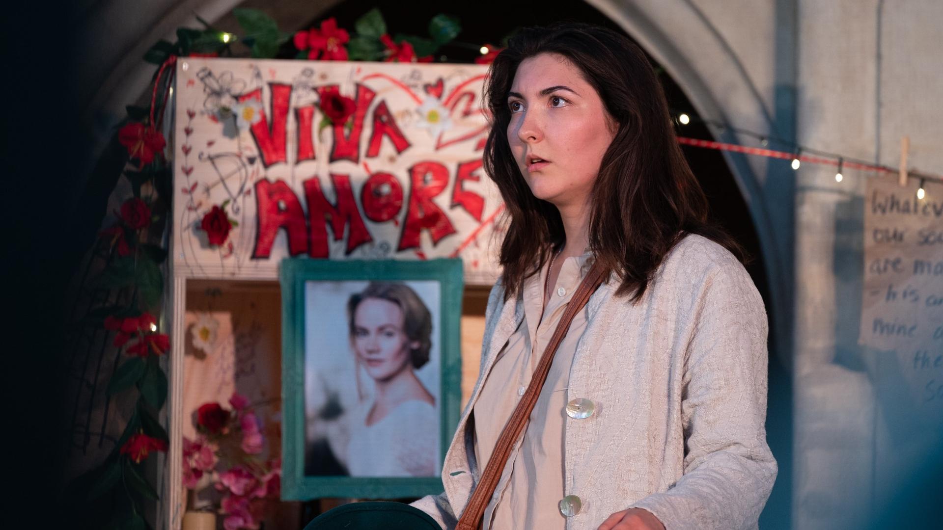 A woman with long brown hair and a light coat stands in front of a decorated memorial made of birch plywood, adorned with flowers, a portrait of another woman, and a sign that reads VIVA AMORE in red graffiti-style letters.