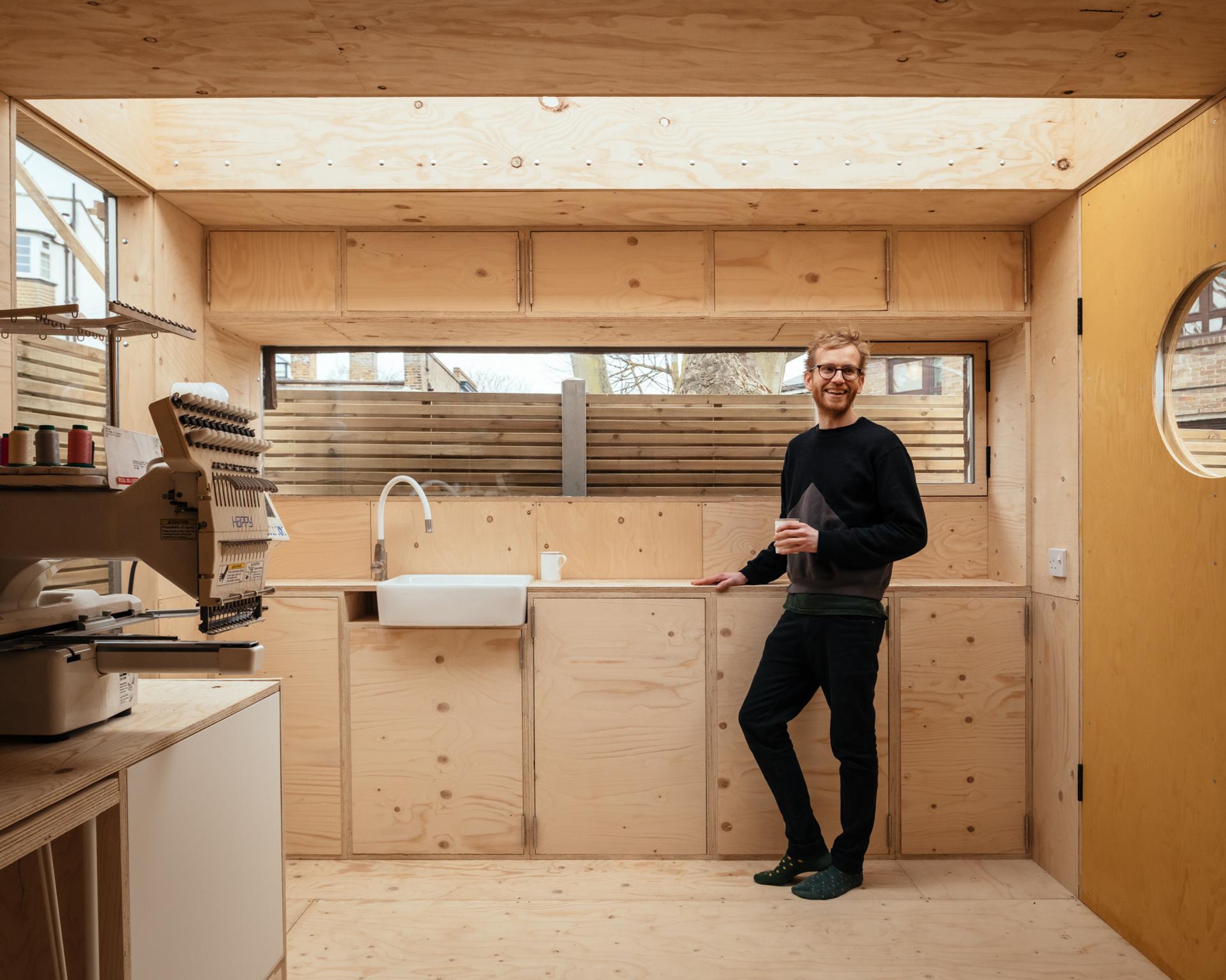 A man stands smiling with a cup in hand in a modern, minimalist kitchen featuring sustainable timber construction, light wood panels, an embroidery machine, a white sink, and large windows letting in natural light.