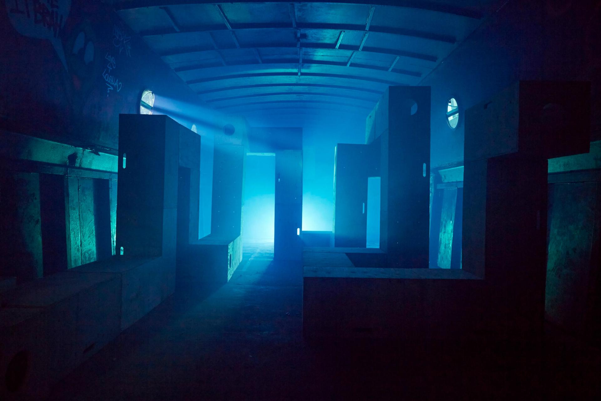 A dramatic, low-light shot of the plywood structures rearranged into L-shaped pillars. The space is bathed in deep blue light with a bright white light source at the far end, creating a silhouette effect and a beam of light cutting through a thick haze or smoke.