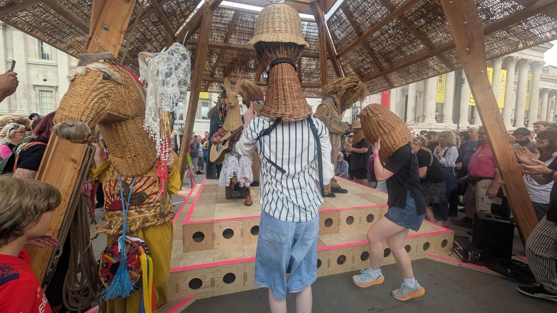 People wearing woven animal head masks perform on a wooden stage outdoors at the National Gallery, surrounded by a lively crowd. The festive event celebrates the Triumph of Art, inviting everyone to Do Your Own Thing with colourful costumes and interactive fun.