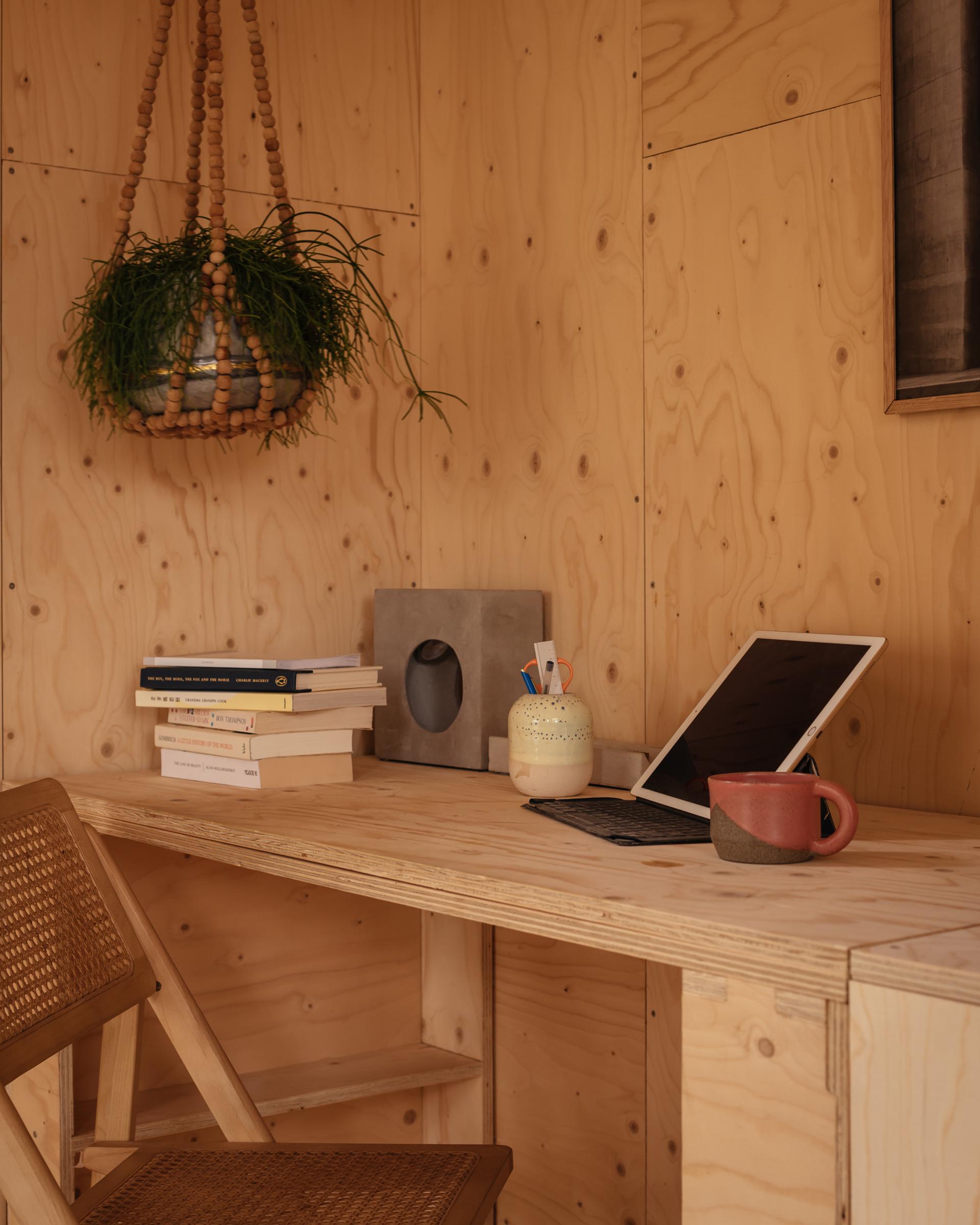 A cosy wooden desk setup featuring sustainable touches: a stack of books, a tablet with keyboard, a cup, a jar holding pens, and a hanging plant in a beaded holder—all set against birch-panelled walls and a woven chair.