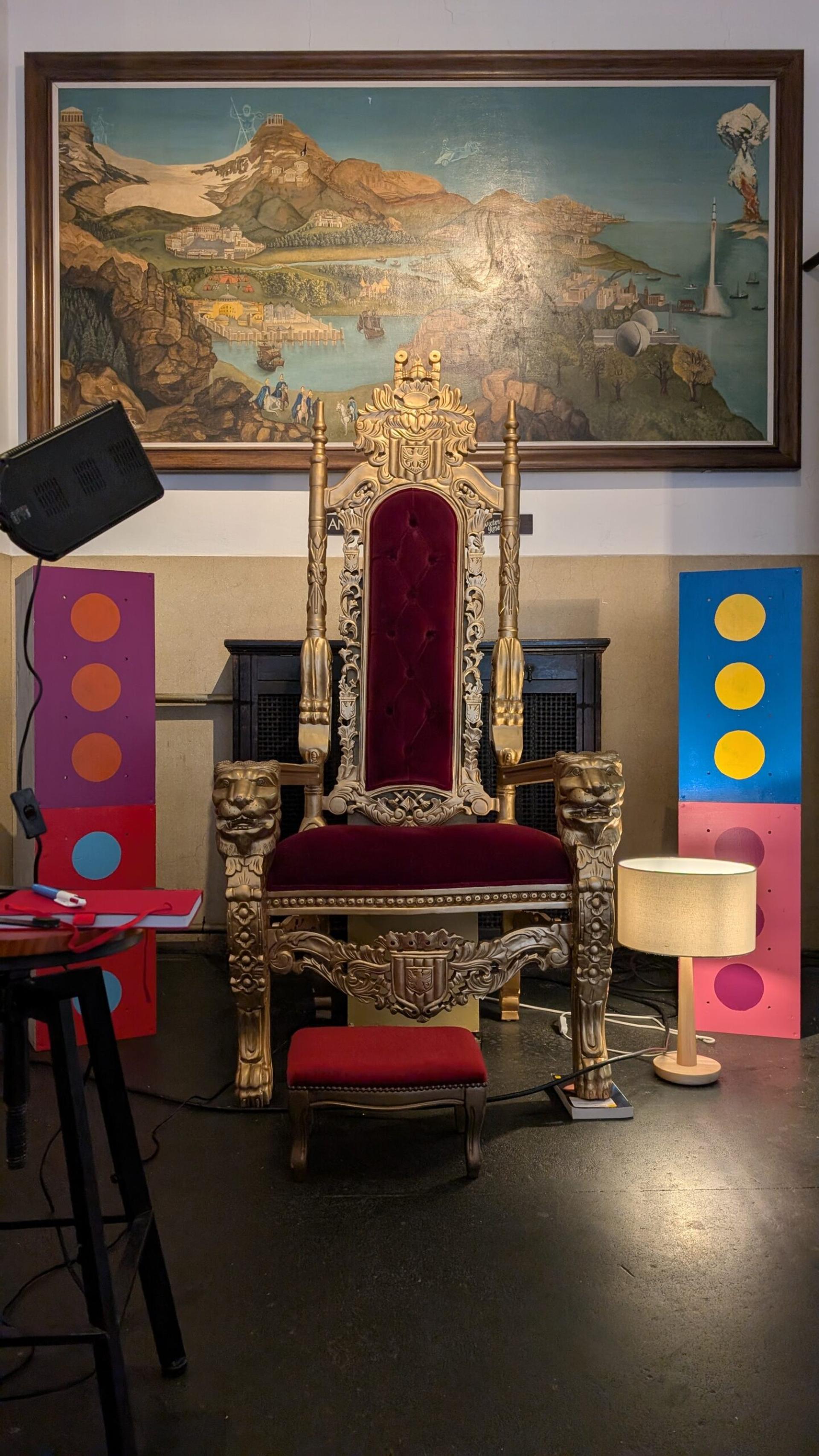 A gold ornate throne with a red velvet cushion sits in the centre, flanked by colourful polka-dot panels and a lamp. A large framed painting of a scenic landscape, crafted from sustainably sourced materials, hangs on the wall behind it.