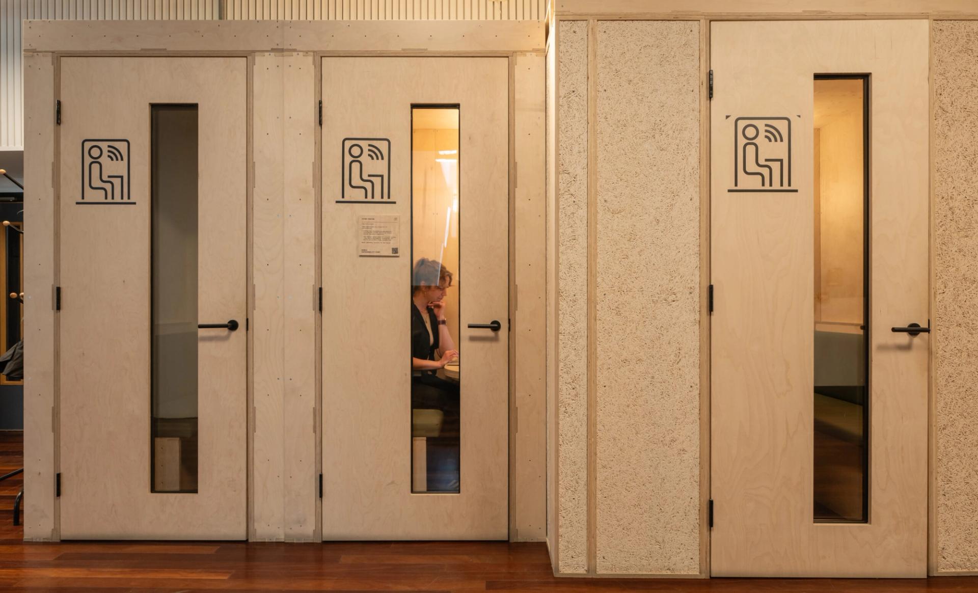 A person sits inside a small, private Meeting Pod with a glass-panelled door, talking on the phone. The pod is one of several lined up in the modern Renold Building, each marked with a conversation icon.