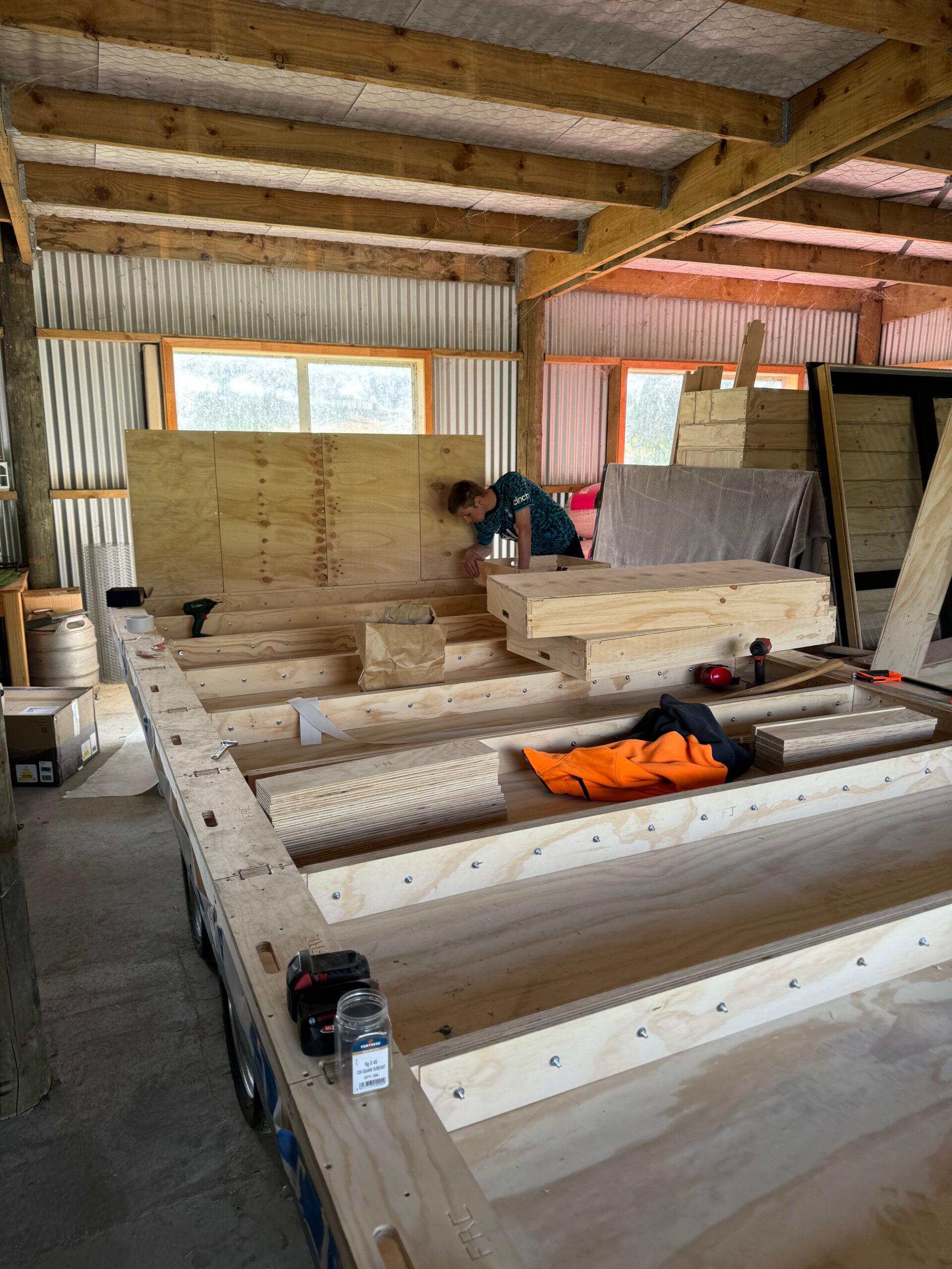 A person works on a spruce wooden structure inside a workshop with exposed beams and corrugated metal walls. Piles of timber, tools, and a high-visibility orange jacket are visible on the workbench.