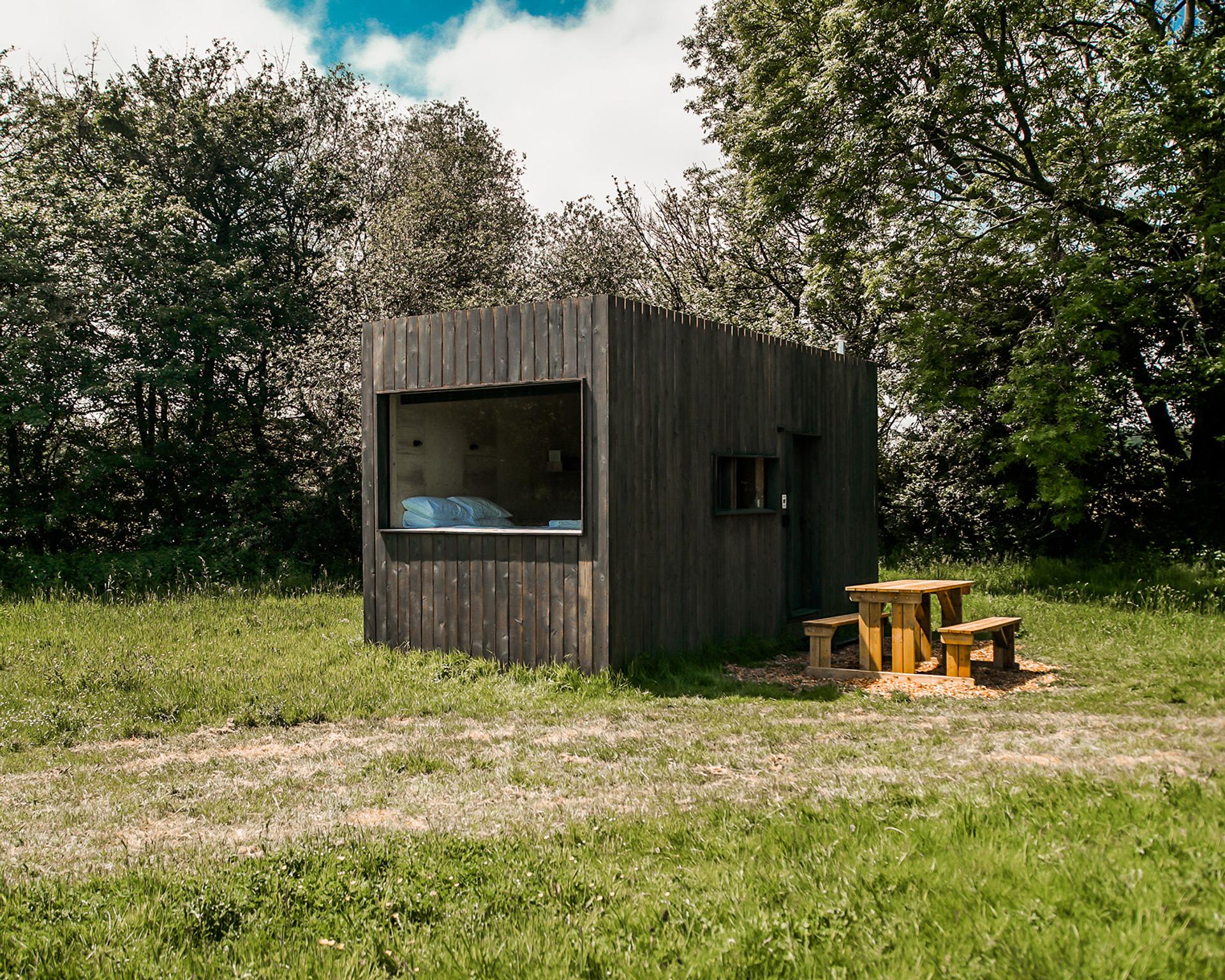 A small modern wooden cabin made from sustainably sourced materials stands in a grassy field, surrounded by green trees. A wooden picnic table with benches sits nearby on a patch of earth under the open sky.