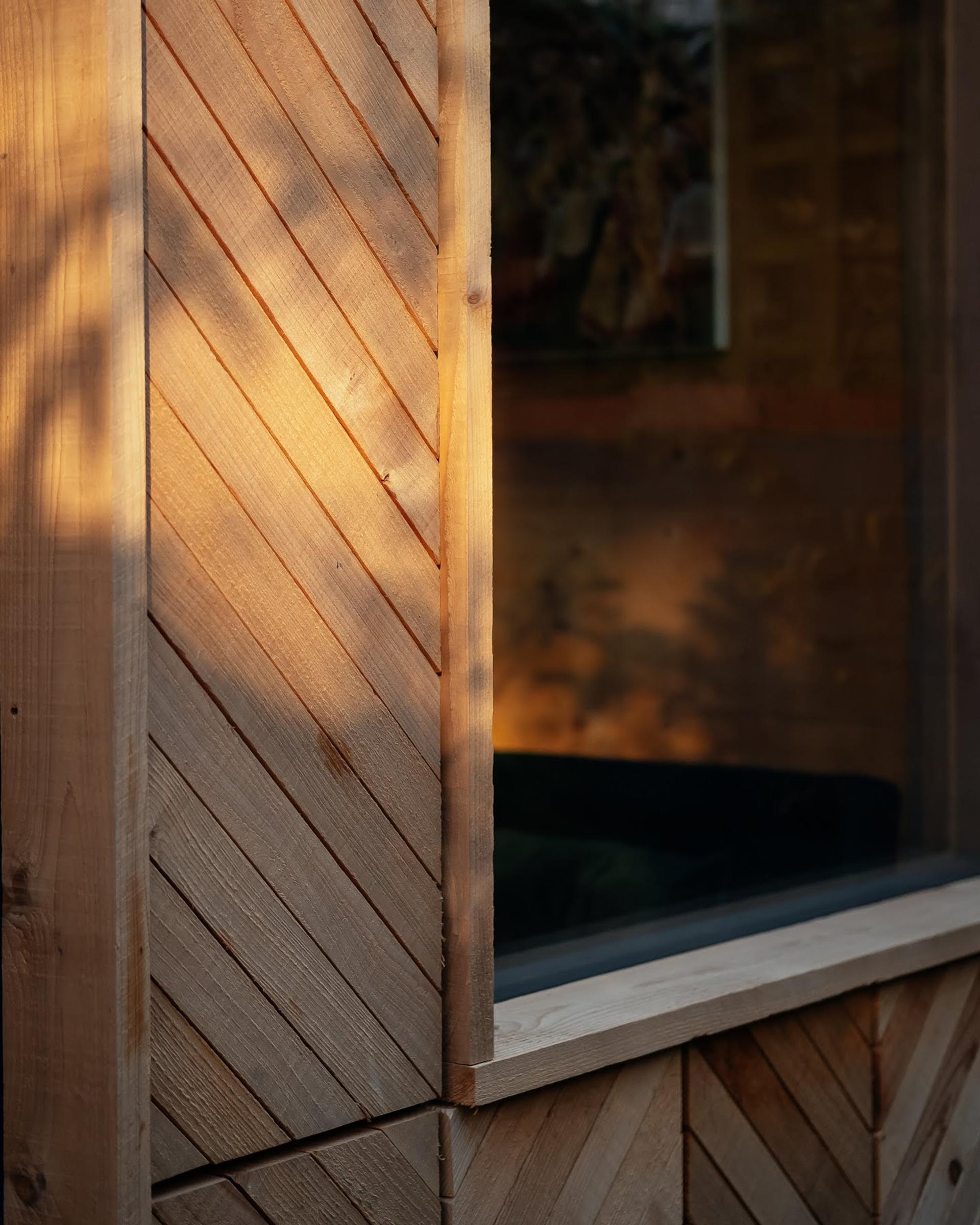 A close-up of a building corner with plywood panels arranged in a chevron pattern, illuminated by warm sunlight next to a window reflecting soft shadows and outdoor scenery, showcasing elegant timber construction.