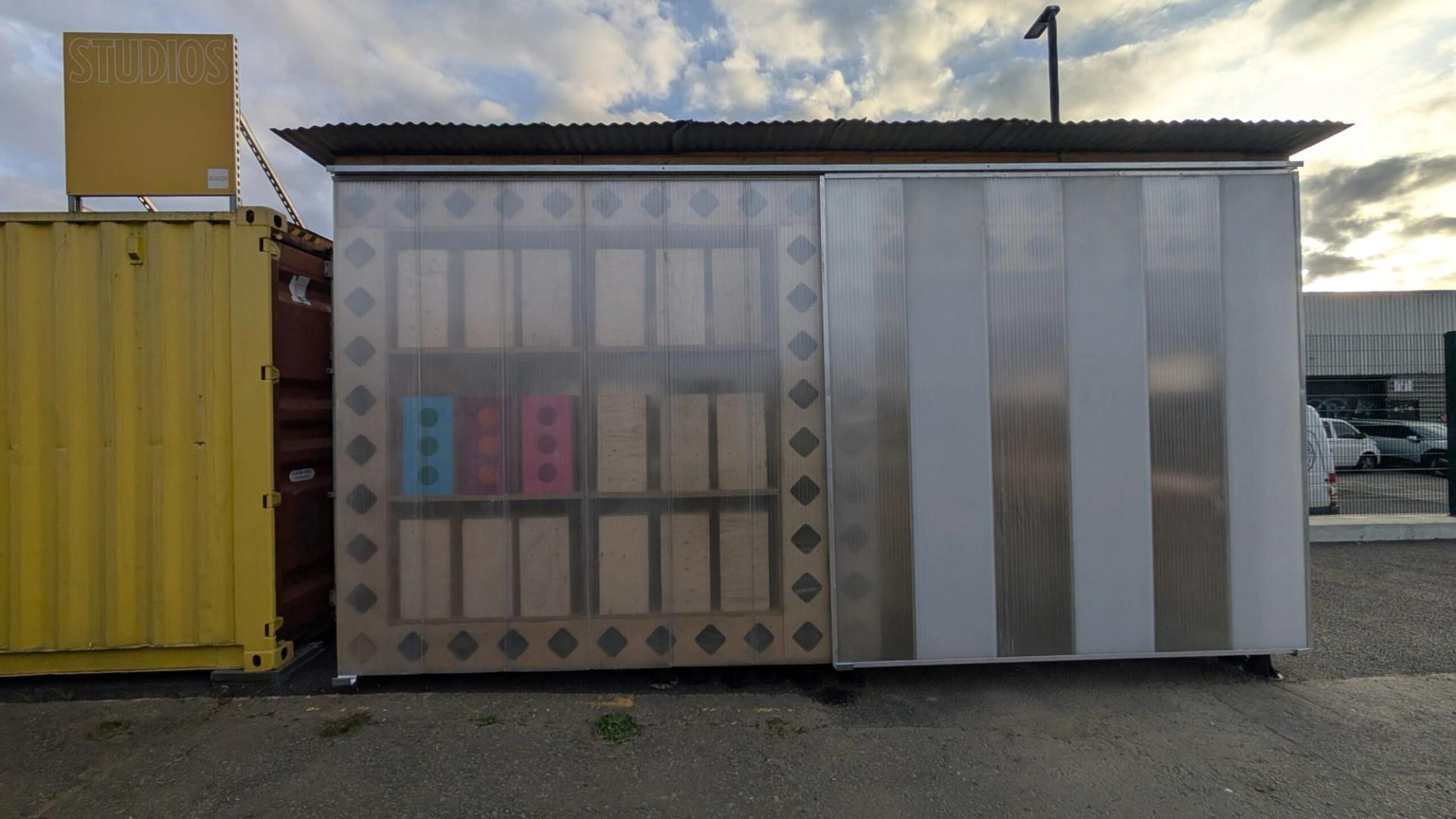 A small U-Build structure with a translucent plastic façade featuring geometric patterns and coloured dots, set between shipping containers in an industrial car park under a cloudy sky.