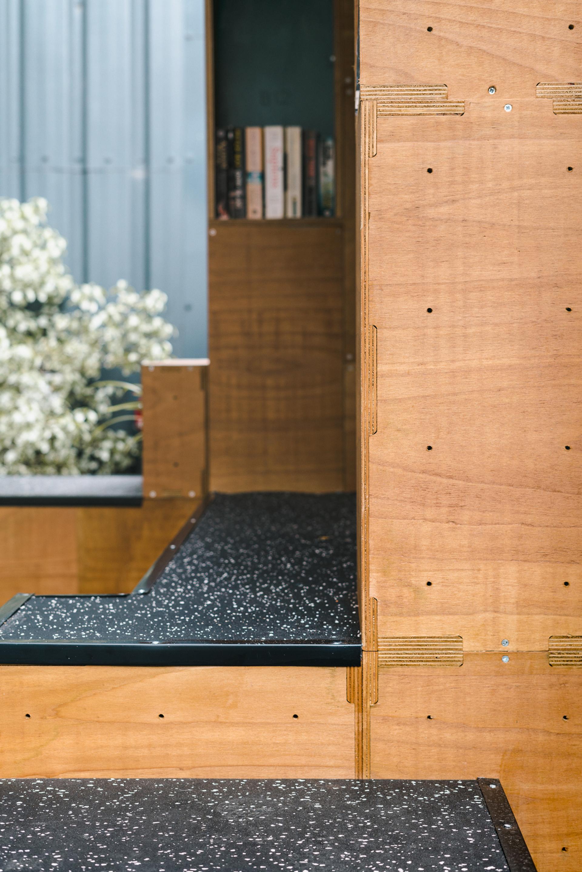 A close-up of a modern wooden staircase with black speckled steps, featuring exposed joints crafted from sustainably sourced materials. A small bookcase with several books is at the top, and blurred white flowers are seen outside a window in the background.