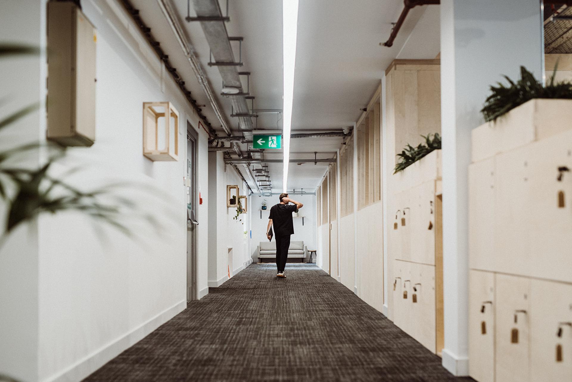 A person walks down a modern corridor with spruce lockers and plants on the right, white walls, modular design elements, and exposed ceiling pipes, heading towards a green exit sign at the end.