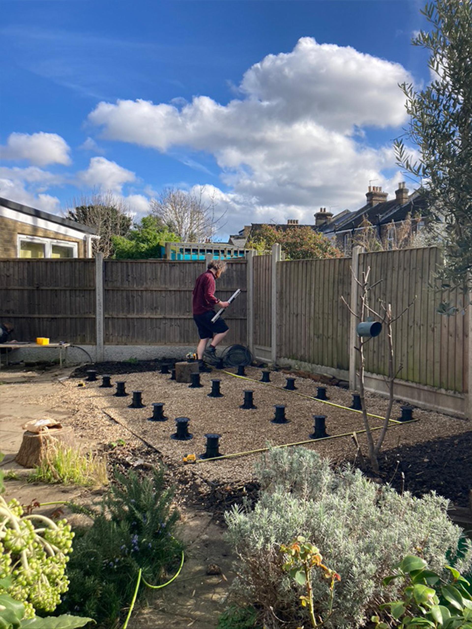 A person stands in a gravel area of a back garden, reading instructions beside rows of black post supports for a sustainable timber decking construction. The sky is partly cloudy, and houses are visible in the background beyond a wooden fence.