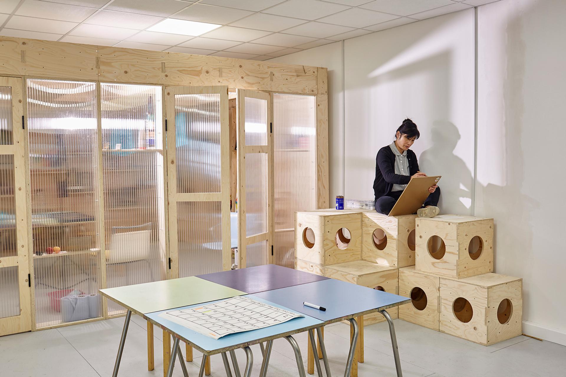 A woman sits on stacked plywood cubes in a bright classroom, writing on a clipboard. Nearby are tables with coloured paper, a calendar, and a marker. The room features timber construction and modular wooden dividers for a modern, organised atmosphere.