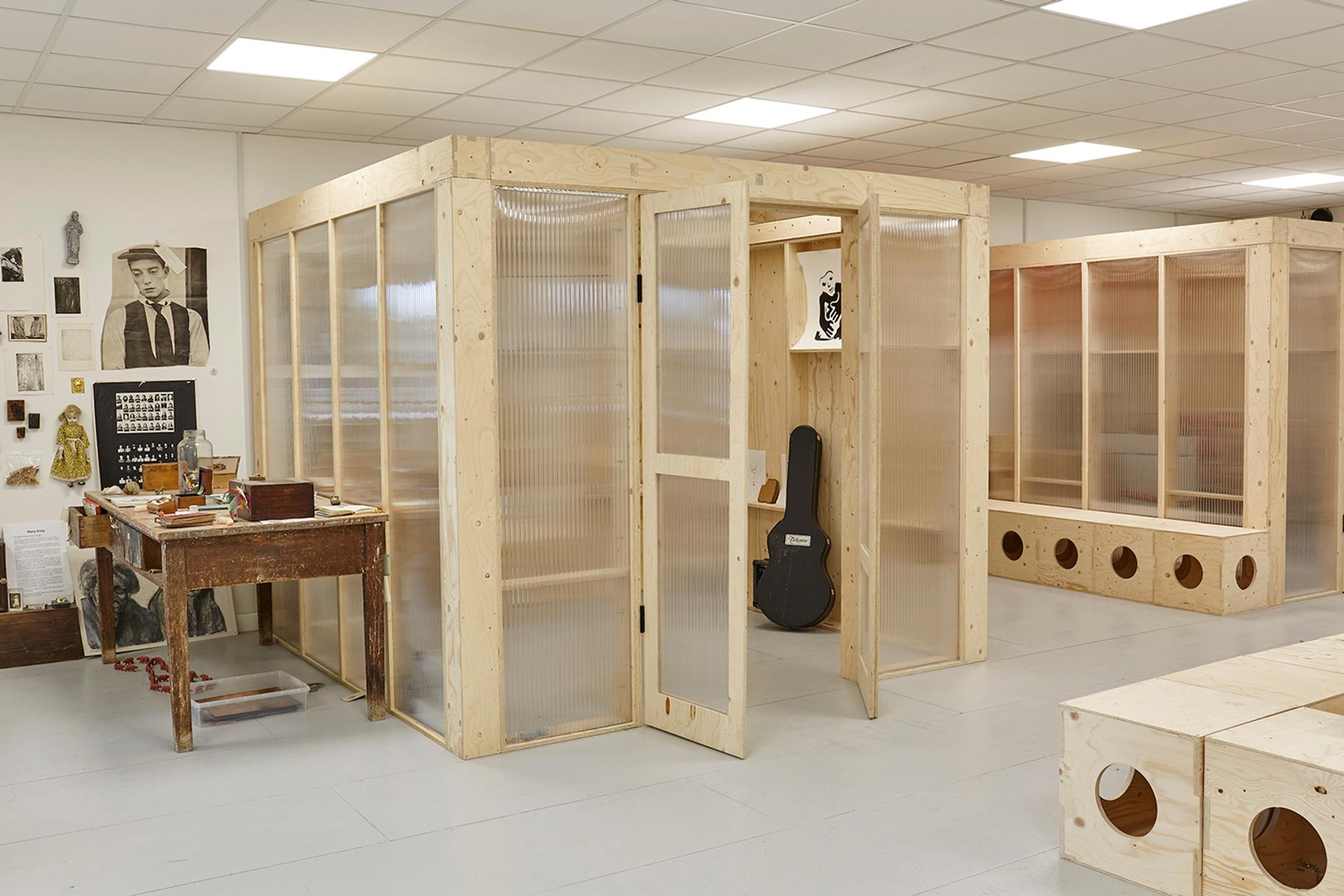 A bright art studio with plywood-framed, transparent partition walls, an open door, a guitar case, and a rustic wooden desk with art supplies; modular artwork and photos are displayed on the walls.